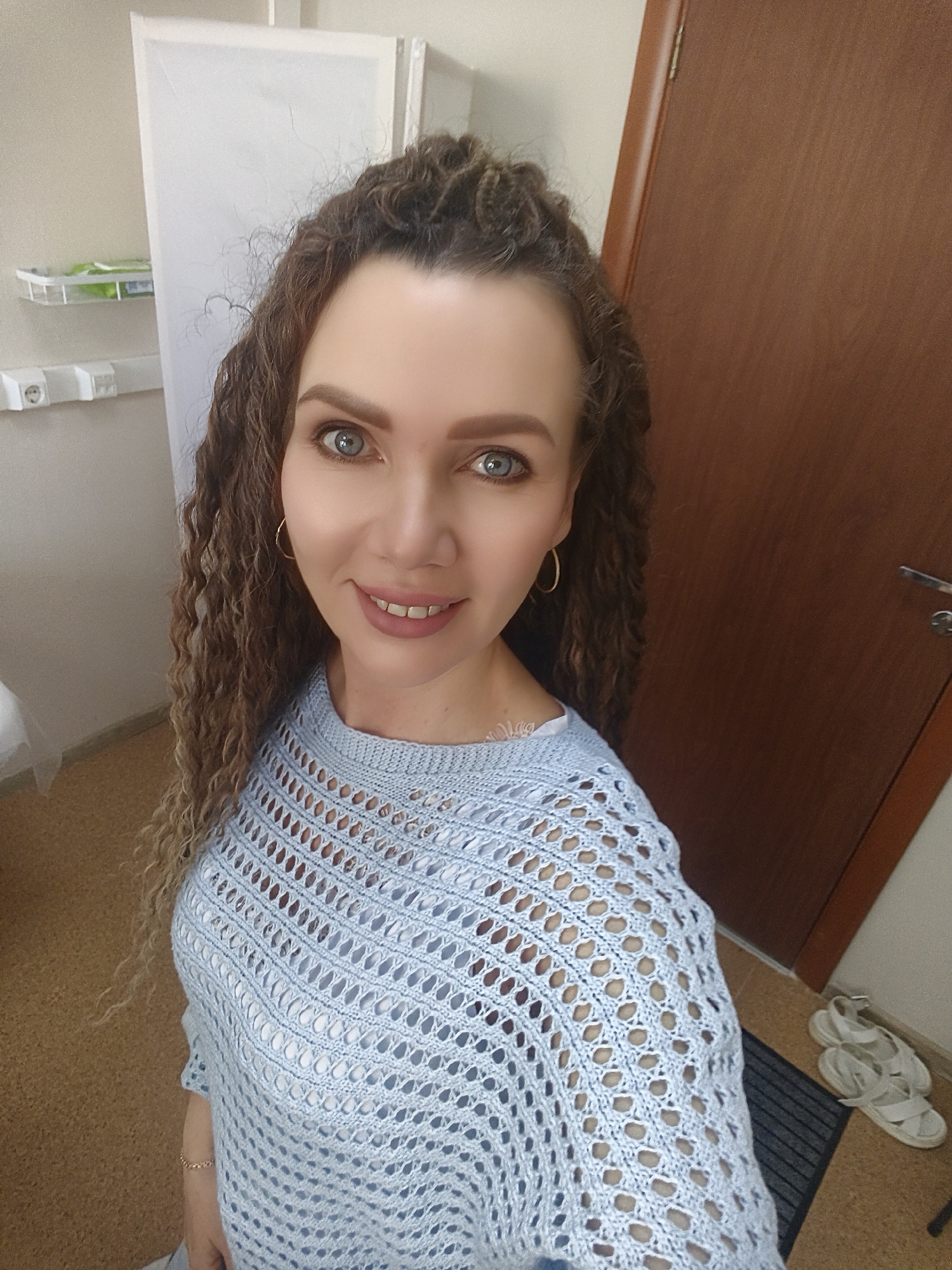 A medium, eye-level shot depicts a woman indoors. Her long, curly, light brown hair cascades around her face. She has light skin, blue eyes, and is smiling slightly. She wears small gold hoop earrings and a light blue, knitted sweater.
In the background, to the left of the woman, is a tall, white, cabinet-like structure. A small shelf containing items is affixed to the wall near the cabinet. To the right, a wooden door with a silver handle is visible. On the floor near the door are white sandals on a small, black mat. The flooring is light brown. The location is in the city of Unknown, Unknown.