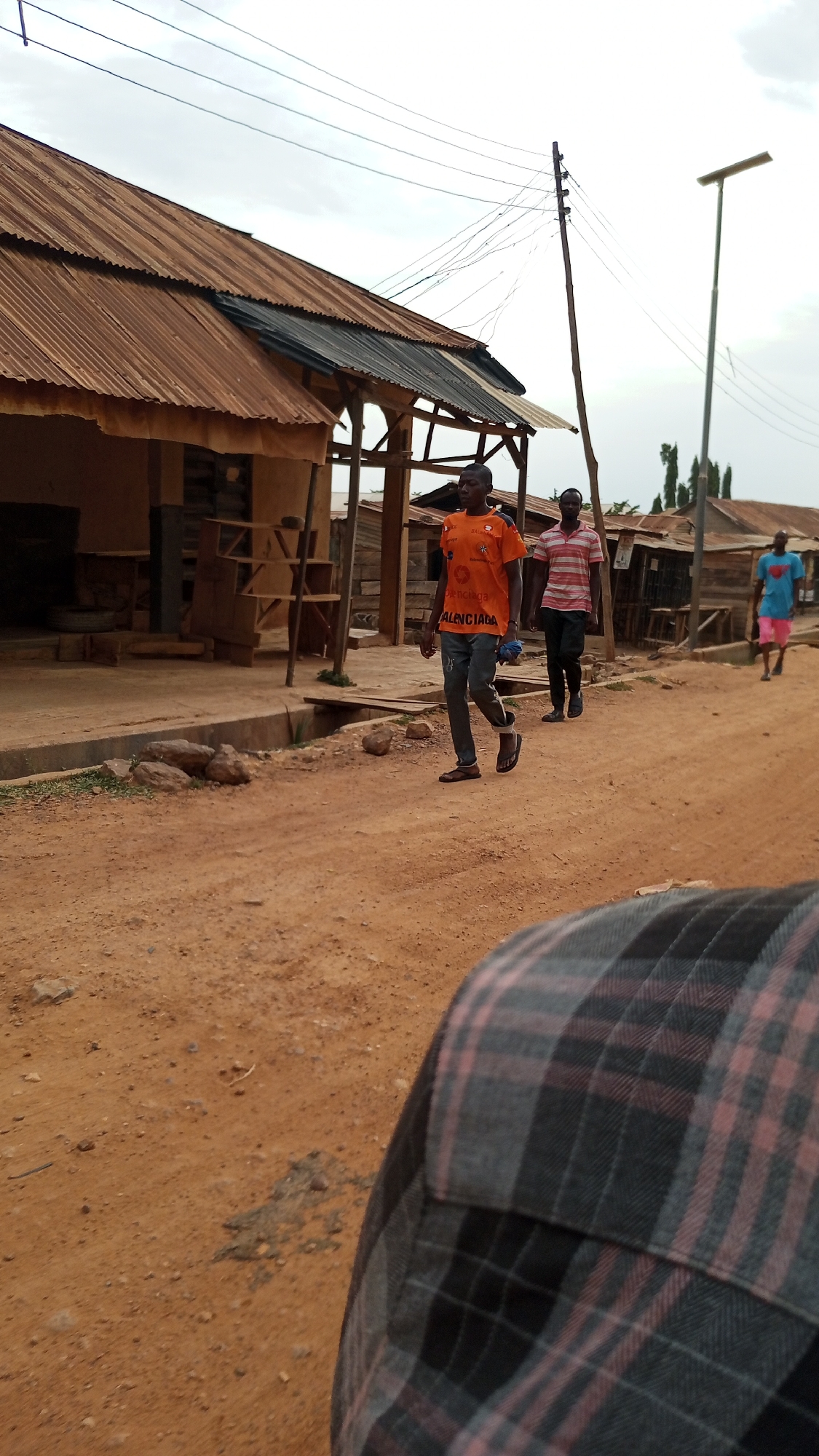 An outdoor scene in Ajaokuta, Nigeria, depicts a dirt road lined with rustic buildings under an overcast sky. Three men are walking along the road from left to right, with the shoulder and back of a fourth person visible in the immediate foreground, wearing a grey, black, and red plaid shirt, suggesting the photo was taken from a close perspective, possibly from a moving vehicle or by someone walking alongside.
The man in the closest full view is wearing a bright orange t-shirt with the word "BALENCIAGA" prominently displayed across the chest in black letters, along with other smaller designs. He wears dark pants, sandals, and holds a small blue object in his left hand. Behind him, another man walks in a striped red and white t-shirt and dark trousers. Further down the road, a third man is visible, wearing a light blue t-shirt and pink shorts.
The buildings on the left side of the road are single-story structures with corrugated metal roofs, showing signs of wear and rust. Exposed wooden supports are visible beneath some eaves. A shallow concrete ditch runs alongside the buildings, separating them from the dirt road, with some rocks and sparse vegetation on its edge. In the background, a tall utility pole with numerous overhead wires stands prominently, and a modern street lamp is also visible further down the road. The time of day appears to be daylight under a cloudy sky. The overall atmosphere suggests a typical day in a semi-rural or developing urban area.