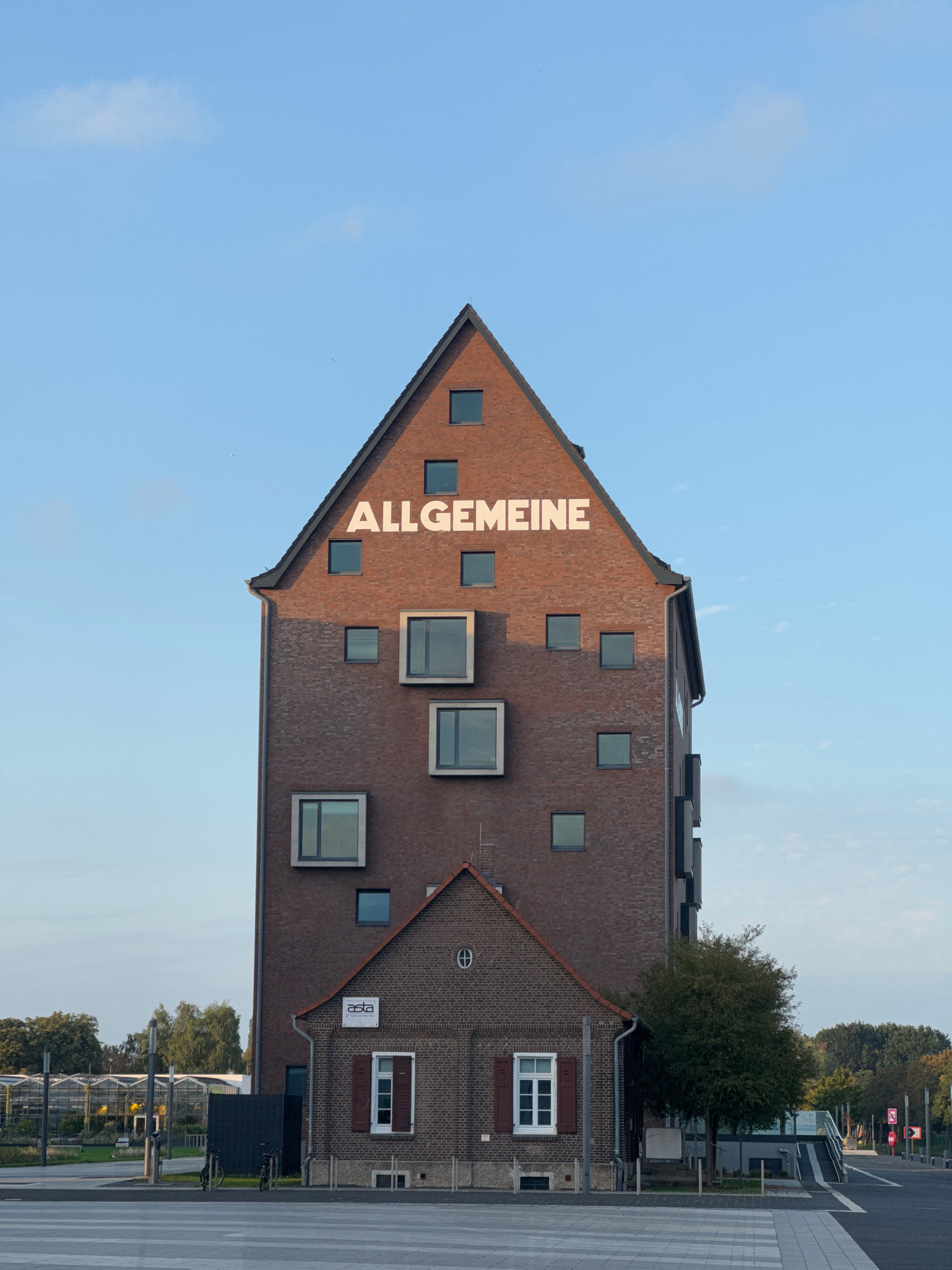 The image shows the imposing brick building of the AStA (Allgemeiner Studierendenausschuss) of the Hochschule Niederrhein in Kleve, Germany. The main structure is a tall, gabled brick building with the word "ALLGEMEINE" inscribed in large white letters on its facade. The building features numerous windows of various sizes and styles, some of which are recessed and appear modern, while others are smaller and more traditional. A smaller, older brick annex is attached to the lower part of the main building. This annex has a gable roof and features a sign with the "asta" logo. The building is situated in an urban environment, with a street in the foreground marked with a pedestrian crossing. Trees and greenery are visible to the right of the building and in the background. The sky is clear and blue with a few wispy clouds. There are no people visible in the image.