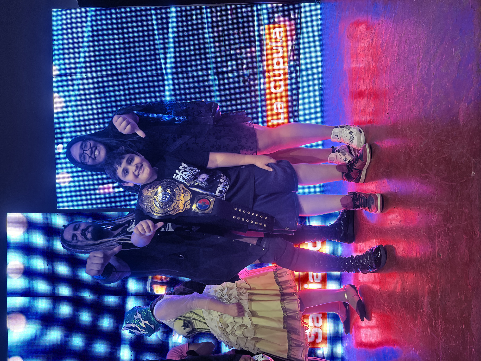A group of four people are standing in front of a large LED screen. The screen displays blue and white abstract patterns, with some words. The floor is made of red and blue flooring.
From top to bottom:
- A person with glasses and a black dress points downward.
- A boy wearing a wrestling belt.
- A man with dreadlocks points upward.
- A person in a yellow dress and a wrestling mask.
The image does not provide explicit information about the location in Arica, Chile. The presence of wrestling belts and masks suggests a wrestling-related event, but the specific venue cannot be determined from the image alone.