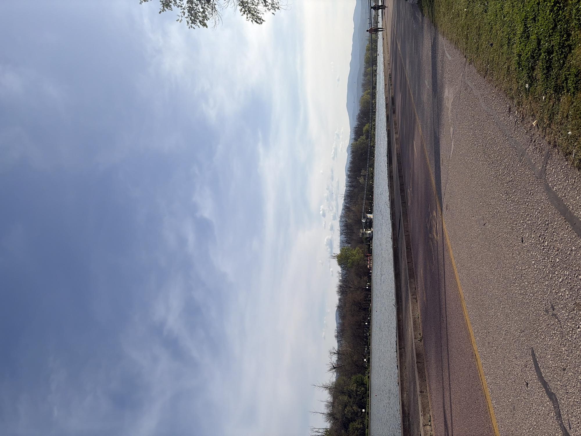 The image shows a landscape with a body of water on the left side, likely a river or lake, bordered by trees. In the distance, there are mountains visible. On the right side is a paved path or road with a yellow line marking. Some grass and vegetation line the edge of the path. In the upper left corner, a small portion of a tree's branches is visible. A person is walking in the distance. The sky is filled with clouds and a light blue color. The location appears to be outside of the city in a natural environment.