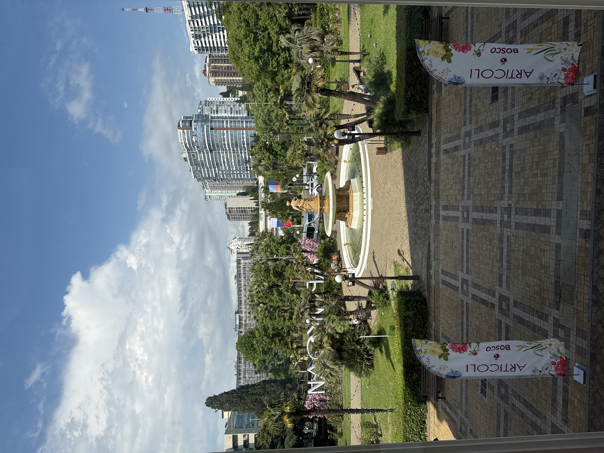 The image depicts a sunlit outdoor scene in Sochi, Russia. The setting is a park-like area with lush green lawns and numerous palm trees, suggesting a warm climate. In the background, modern high-rise buildings with distinctive architectural styles are visible, hinting at an urban environment adjacent to the park.
The foreground features a pathway paved with light-colored bricks, leading towards a decorative fountain. Two banners are prominently displayed on poles, each featuring floral designs and text. One banner reads "ARTICOLI BOSCO" in a vertical orientation, with the text "ARTICOLI" in larger font than "BOSCO". The other banner, partially obscured, appears to have similar text and design.
A large sculptural installation with the Cyrillic letters "ЛЕТО НЕ ДВИЖИМОСТЬ" (Summer Real Estate) is visible in the middle ground, adorned with colorful flags. This text suggests a promotional event or a permanent installation related to summer or real estate. The presence of benches and lampposts indicates a public space designed for leisure and recreation.
The weather appears to be clear and sunny, with blue skies and scattered white clouds. The time of day is likely daytime, given the bright sunlight casting shadows. No people are clearly visible in the frame, contributing to a sense of peacefulness or perhaps early morning. The overall impression is one of a well-maintained public space in a resort city.