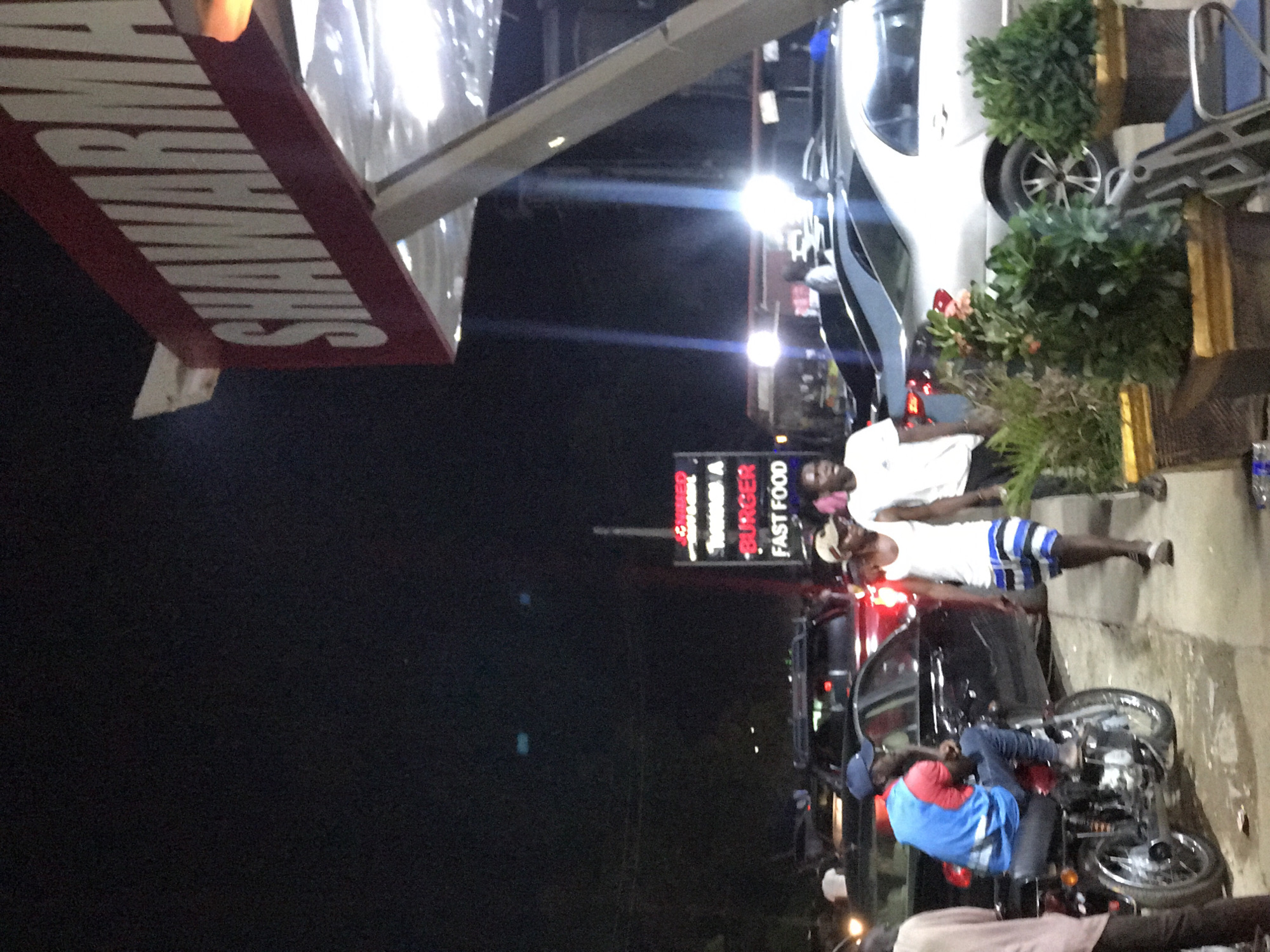 The image depicts a street scene at night in Gwarinpa, Nigeria. A sign is visible for a fast food restaurant called "Burger." Two people are standing on the sidewalk near a parked car, and one of them is wearing a white shirt and striped shorts. A person wearing a blue shirt and jeans is sitting on a motorcycle. There are potted plants near a white car in the background.