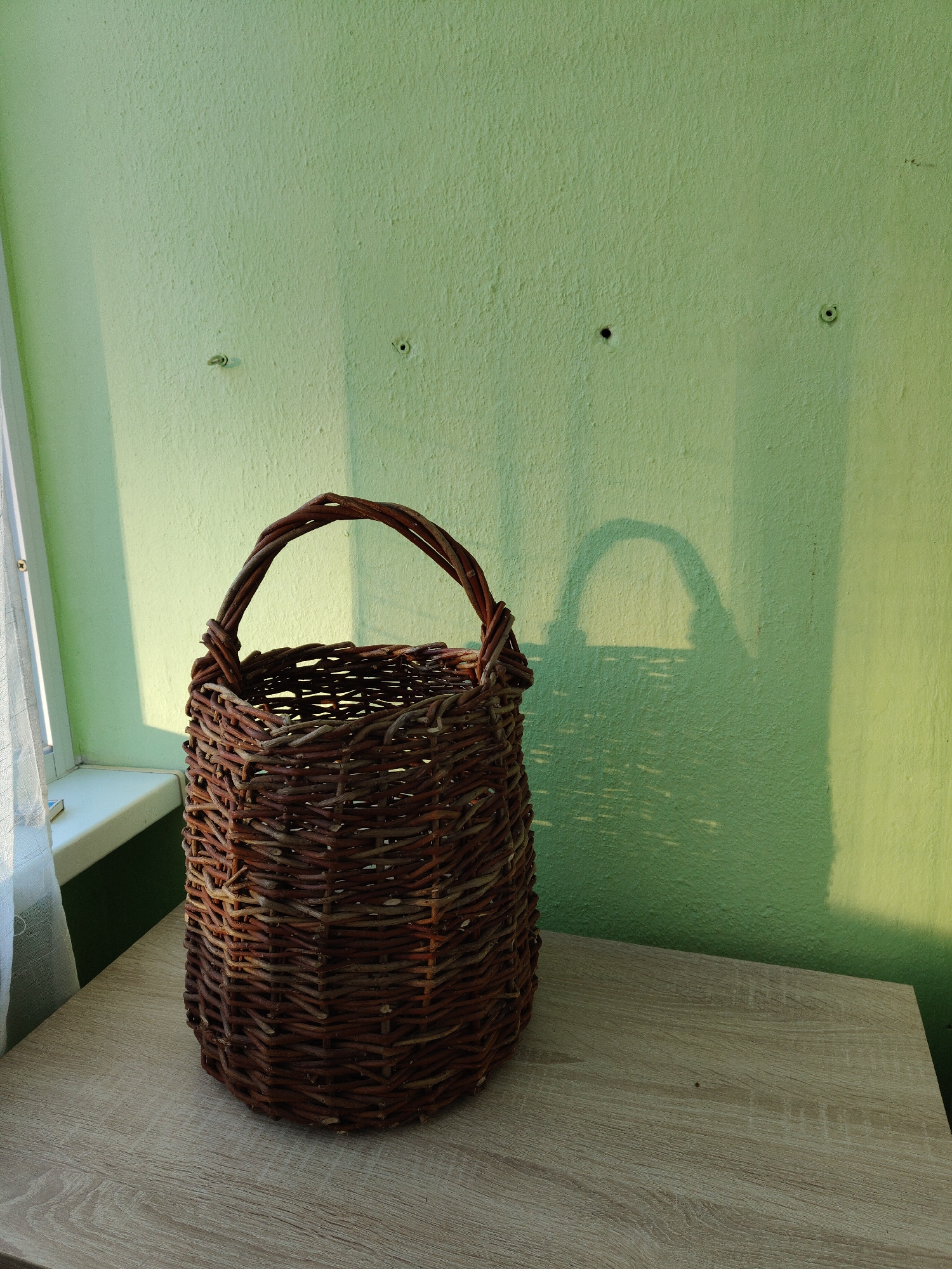 A brown woven basket sits on a light brown laminate surface, positioned against a light green textured wall. A shadow of the basket is cast onto the wall. The basket has a handle. The wall features three wall anchors. To the left of the basket, a window is partially visible, with a white sheer curtain. The location is in the city of Unknown, Unknown.