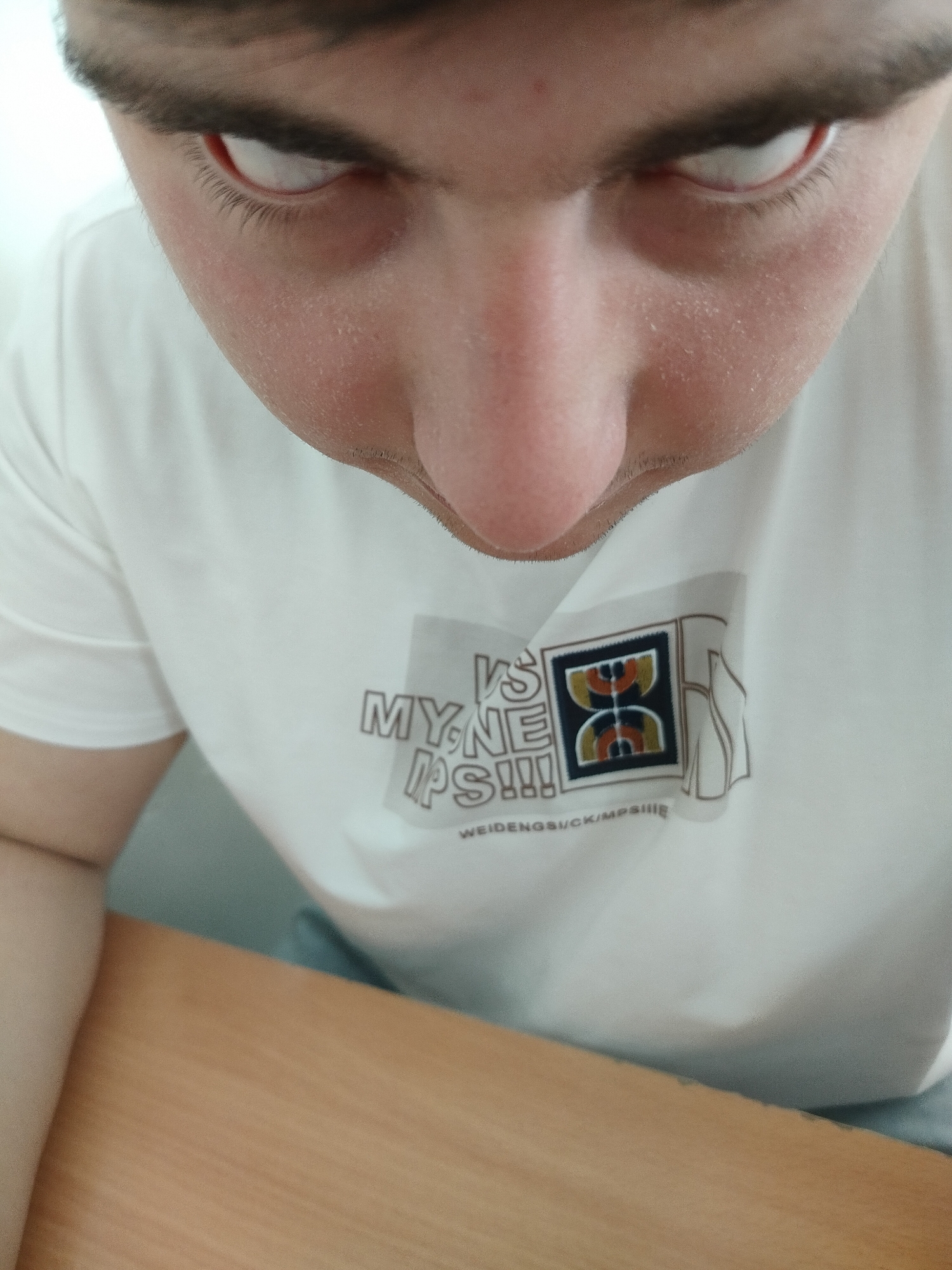 A close-up, high-angle shot depicts a person's face from the forehead down to the upper chest. The person has fair skin, dark hair, and red eyes. They are wearing a white t-shirt with a graphic design that includes the words "MY.NE VS PS!!!" and "WEIDENGSUCK/MPSIII," along with a graphic with a geometric design. A wooden table is visible in the lower part of the frame. There are no indicators of Yerevan, Armenia, or specific locations.