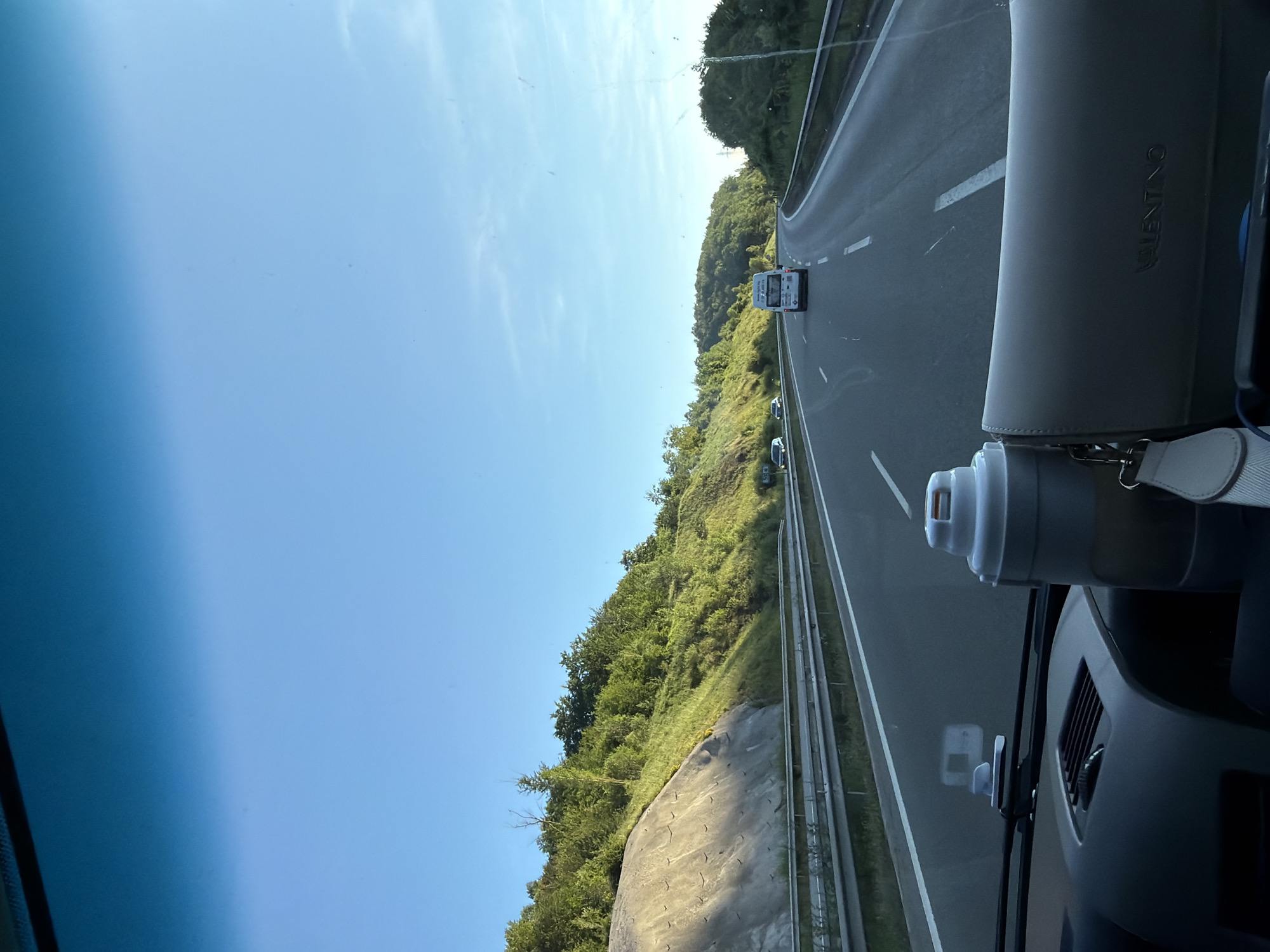 The image is taken from inside a vehicle, looking forward through the windshield at a highway. The sky is clear and blue. On the right side of the highway, there are trees and foliage covering rolling hills. On the left side, a steep, rocky embankment is visible, also covered with greenery. Several vehicles are on the highway, including a white van and a few cars. In the foreground, on the dashboard, a white insulated cup and a light-colored purse are visible. The image is consistent with a scene on a highway in a rural or semi-rural area, such as the outskirts of Malo Buchino, Bulgaria, during daylight hours.