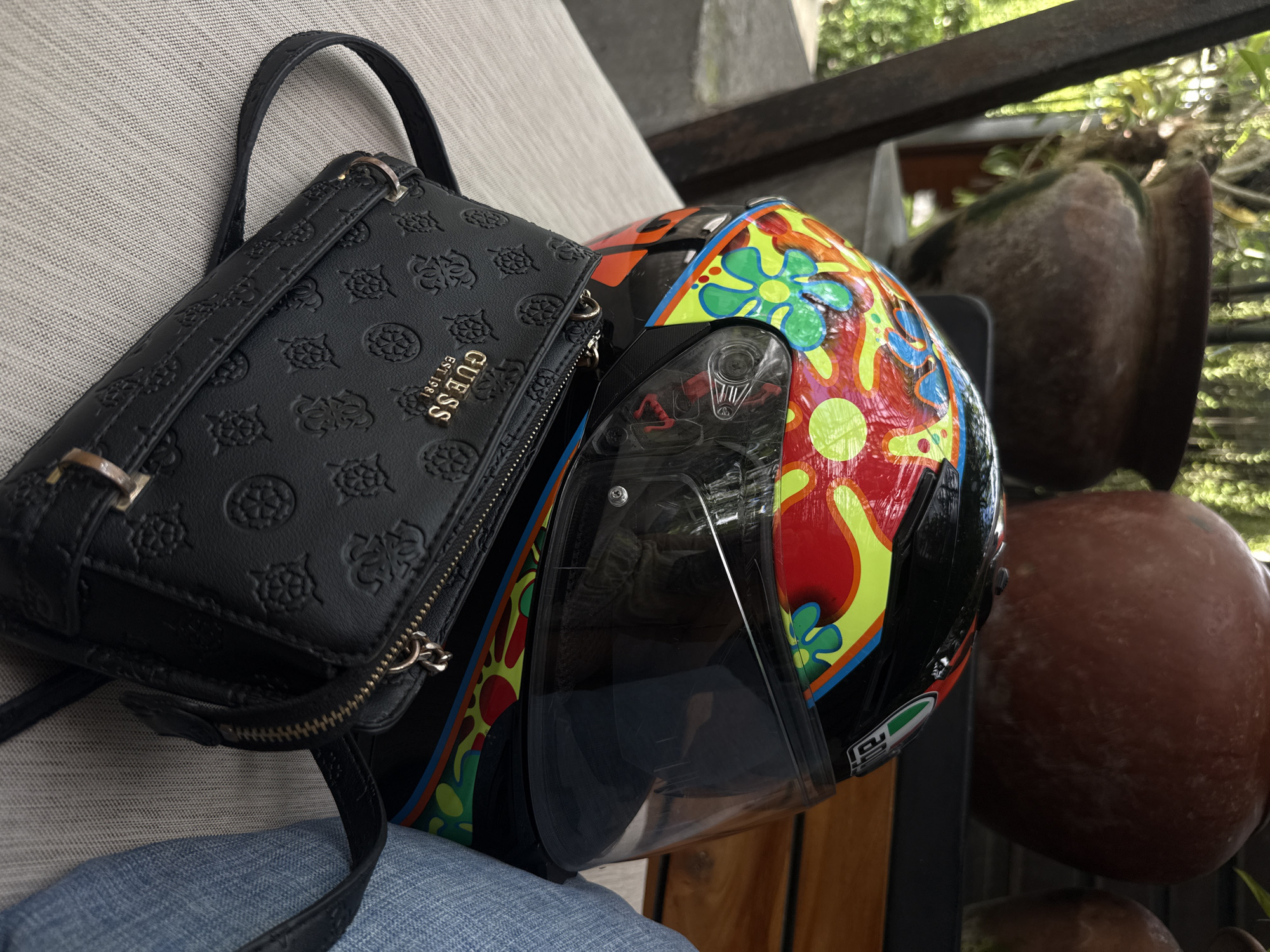 The scene captures a casual setup, likely outdoors, possibly in North Kuta, Indonesia, though not definitively proven by the image itself. A black Guess handbag rests adjacent to a colorful motorcycle helmet. The helmet, predominantly black, features a vibrant design of stylized flowers in shades of red, yellow, green, and blue. The helmet's clear visor is partially open. The objects are placed on a textured, light-colored surface, possibly a cushion or seating area. Background elements include partially visible potted plants, suggesting a patio or garden setting, and wooden architectural details. A small patch of blue fabric is visible beneath the objects. The image is well-lit, allowing for clear identification of the items and their immediate surroundings. There is no evidence of any people in the picture.