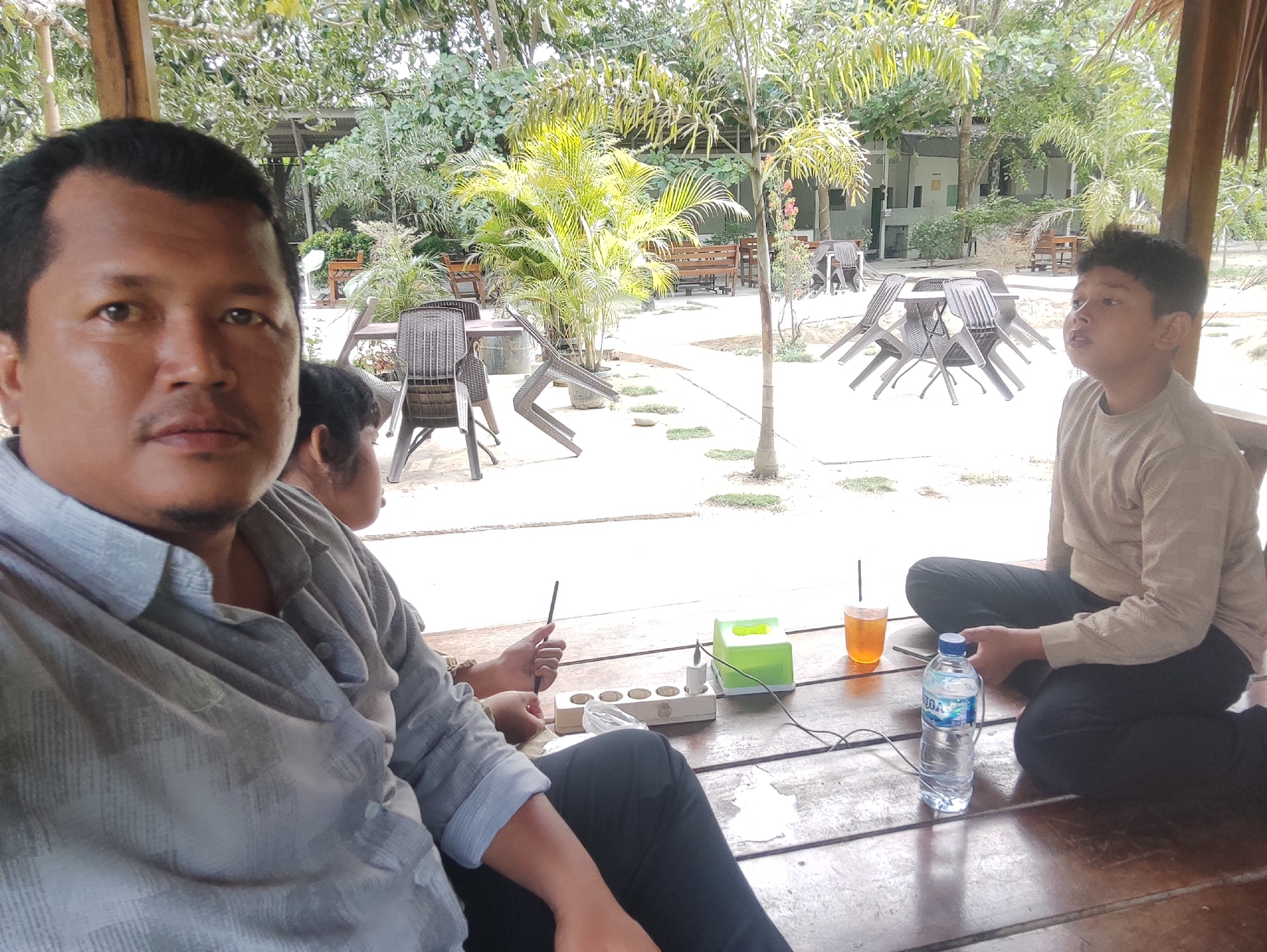 This image captures a relaxed outdoor scene in Keudealue Ie Puteh, Indonesia, likely at a cafe, restaurant, or resort. The perspective is a close-up selfie or a shot taken from very close to the foreground subject.
In the immediate foreground, on the left, a man with short dark hair and a mustache looks directly at the camera with a neutral expression. He is wearing a light gray patterned long-sleeved shirt and is seated on a wooden floor. Partially obscured to his right, another individual's hand is visible, holding a thin black object, possibly a pen or stylus. On the wooden surface in front of them are a white power strip with multiple sockets, a green tissue box, and a glass of iced tea with a straw. A plastic bottle with the word "AQUA" clearly visible on its label also sits on the floor.
To the right, a boy or young man sits cross-legged on the wooden floor, looking upwards and to his right with a contemplative or focused expression. He has dark, spiky hair and is wearing a light brown long-sleeved shirt and dark pants.
The background reveals a spacious open-air area under a partial thatched or wooden roof structure. Numerous outdoor dining tables and dark brown rattan-style chairs are arranged across a light-colored ground surface. The environment is lush with tropical foliage, including various trees, palm-like plants, and shrubs. Several wooden benches and distant buildings are also visible. The bright, even lighting suggests it is daytime, likely a sunny morning or afternoon. The overall atmosphere is one of leisure and tranquility.