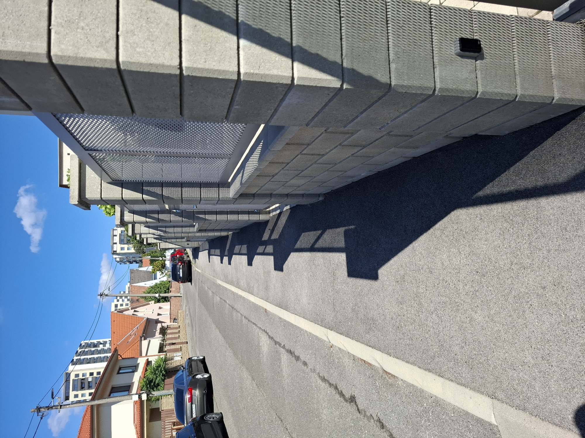 The image depicts a residential street scene in Bratislava, Slovakia, on a clear, sunny day. In the foreground, a modern grey concrete wall with a metal mesh infill runs along the right side of the frame. A black security or access control device is mounted on the rightmost pillar of this wall. To the left of the wall is a paved sidewalk, and further left is a paved street. Several cars are parked on the street and in driveways: a dark grey Audi sedan, a dark SUV, and a red hatchback are visible. In the background, residential houses with orange tiled roofs are situated, and beyond them, modern apartment buildings rise against the blue sky with scattered white clouds. Power lines and poles are also present in the background. The lighting suggests it is daytime, with distinct shadows cast by the wall and trees.