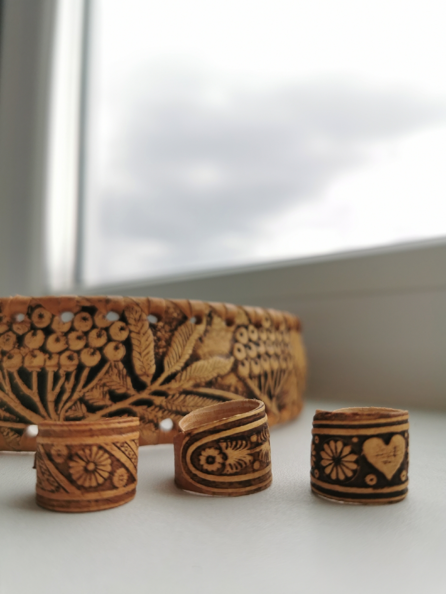 The image shows three small, cylindrical objects crafted from wood, possibly birch bark. The objects are adorned with engraved patterns, including floral motifs and a heart. A larger, shallow wooden basket with similar engraved designs is positioned behind them. The scene is set on a white surface, likely a windowsill, with a blurry window and overcast sky in the background. The location is Unknown, Unknown.