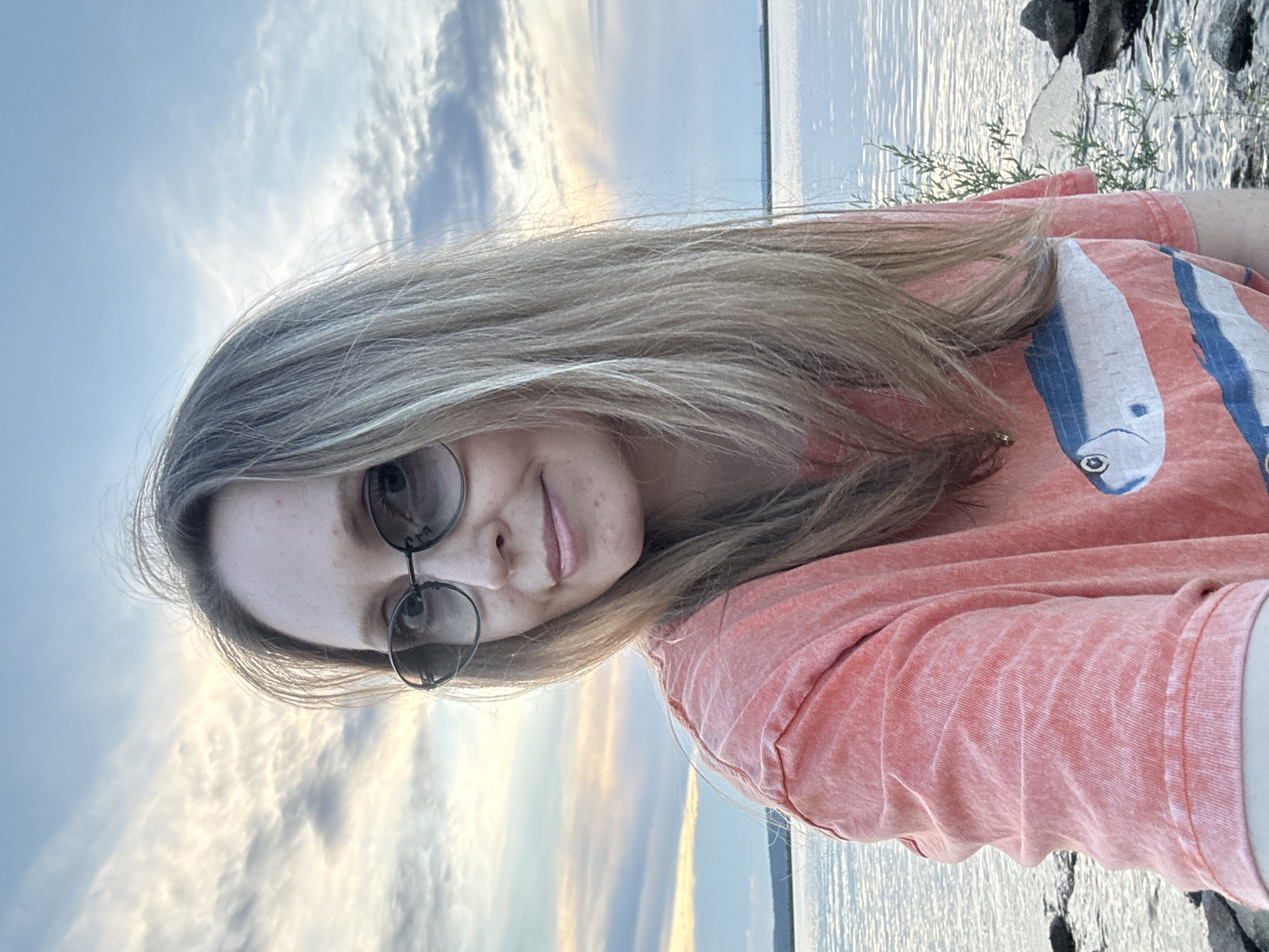 A woman is taking a selfie at a body of water, likely a river or lake. She is wearing sunglasses and a coral-colored t-shirt with a graphic print of two fish. The background shows a sky with scattered clouds and the distant shoreline. The scene appears to be outdoors, possibly at dusk. The location is specified as Chaikovsky, Russia.