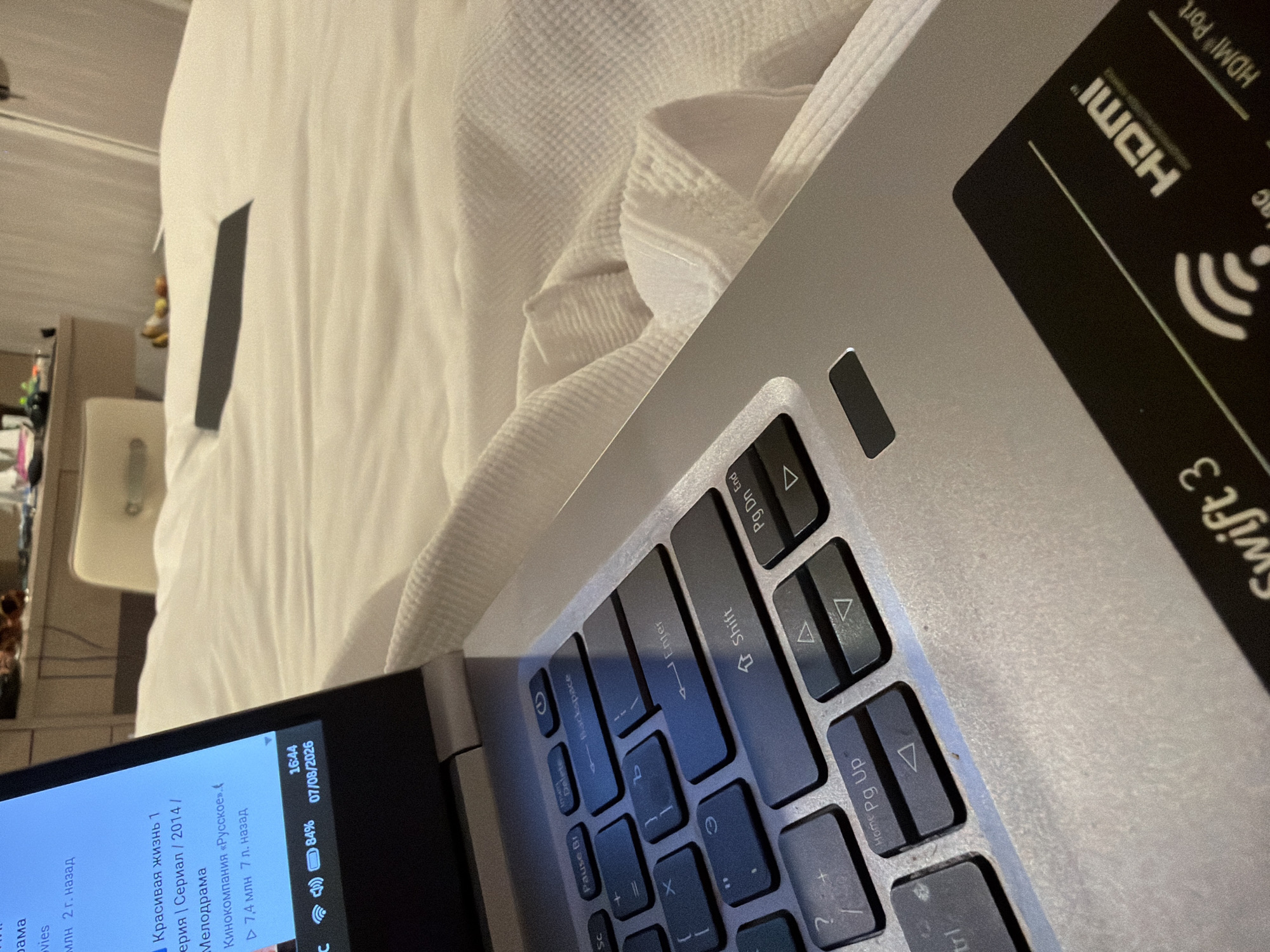 The image shows a close-up of a silver laptop resting on a white, textured fabric, likely a bedspread. The laptop is open, with the screen displaying a list of movie titles in Russian, including "Красивая жизнь 1" (Beautiful Life 1) and "Мелодрама" (Melodrama). The keyboard is partially visible, showing keys like "Enter", "Shift", and navigation arrows. In the bottom right corner of the laptop, a sticker with "HDMI" branding is present. The overall environment appears to be an indoor setting, possibly a bedroom, with soft lighting. There are no people visible in the image.