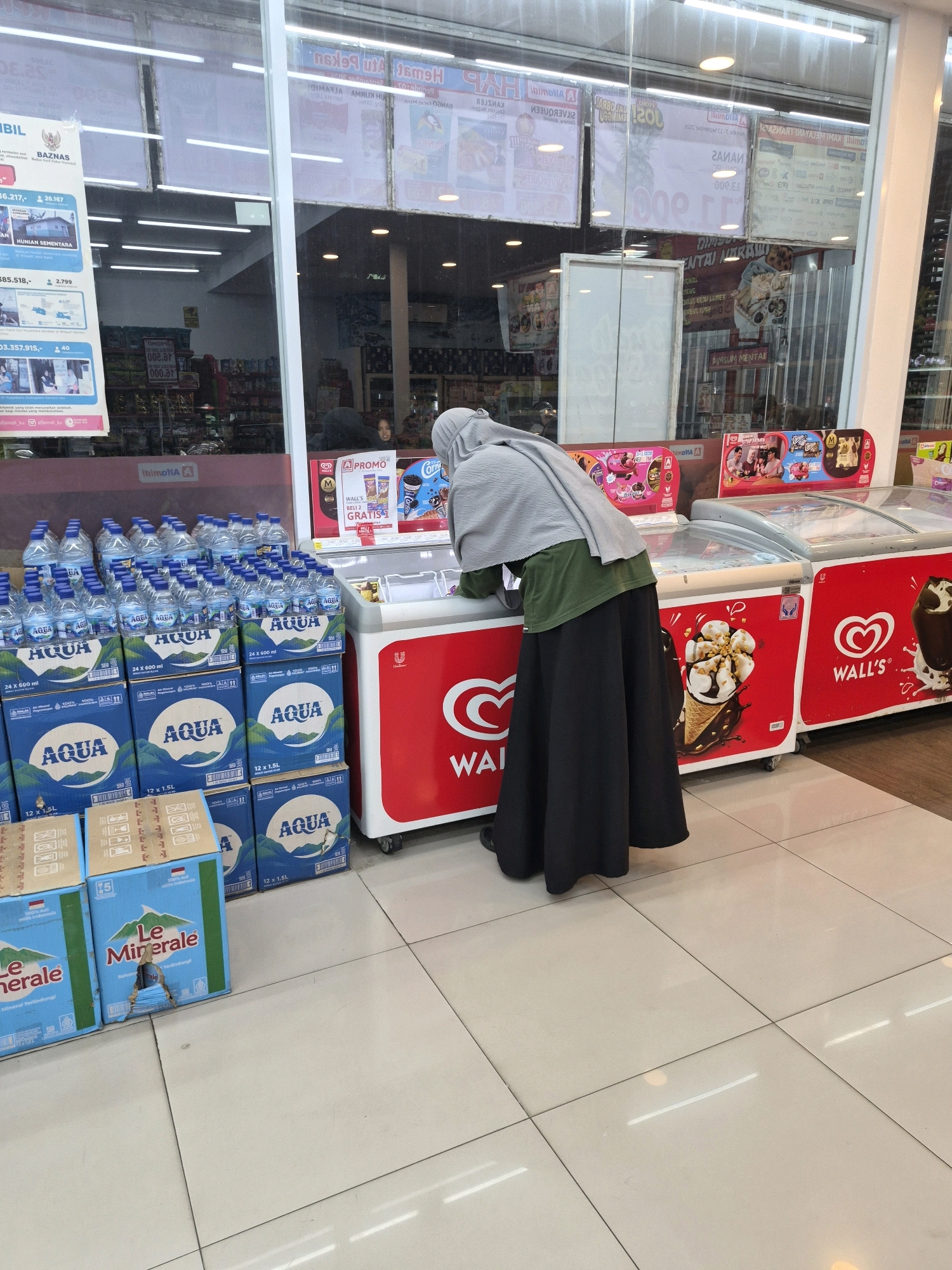 The image depicts the interior of a convenience store in Purworejo, Indonesia, with a focus on a person wearing a hijab and a long black skirt, reaching into a chest freezer. In the foreground, stacks of bottled water from "Aqua" and "Le Minerale" are visible, indicating a retail environment. The freezer itself is branded with "Wall's" ice cream advertisements, suggesting it is a commercial freezer. The store appears to be well-lit with fluorescent lighting, reflecting off the tiled floor. Behind the freezer, shelves stocked with various products are partially visible, suggesting a typical convenience store setup. The overall scene conveys a sense of everyday commerce.