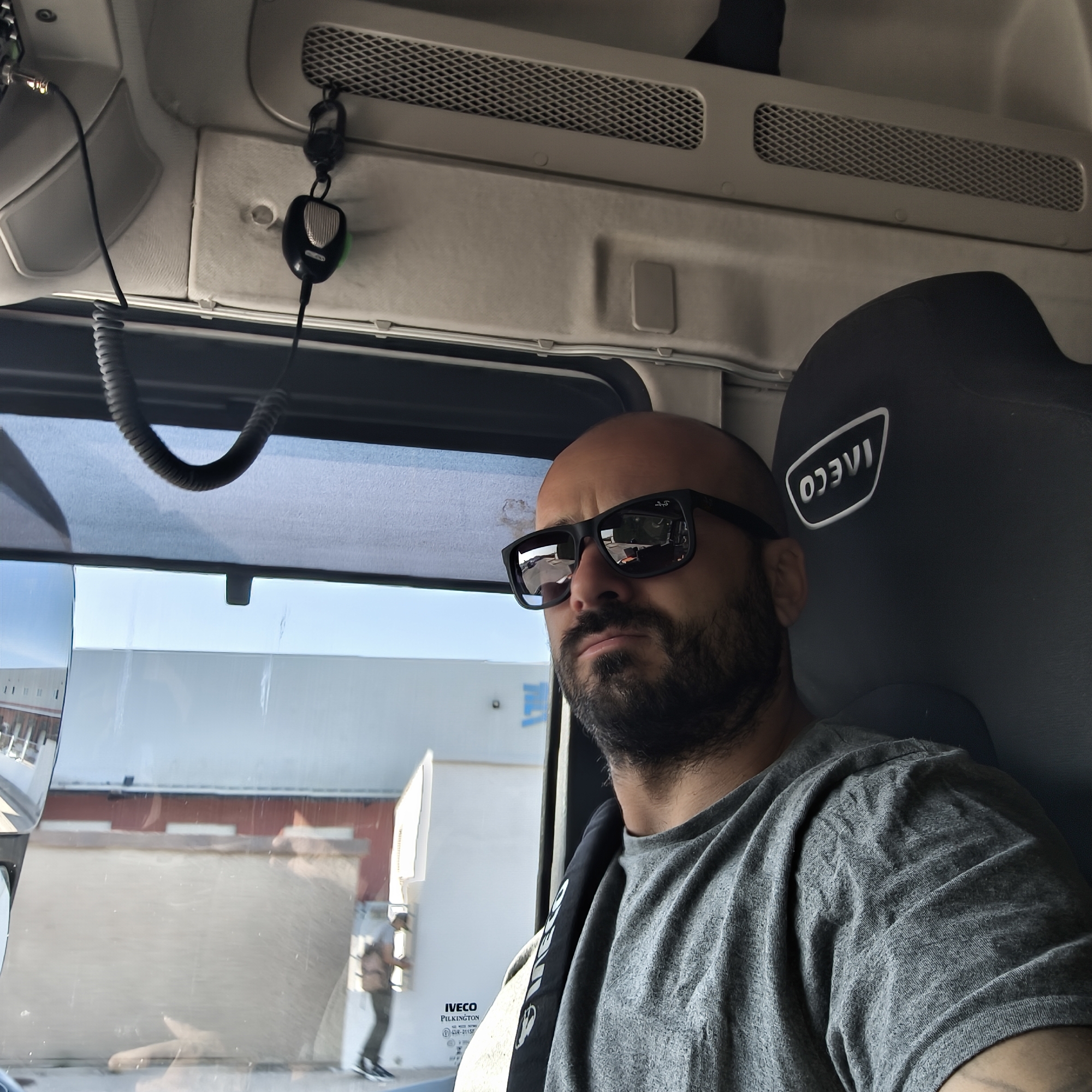 A man with a bald head and a full beard, wearing dark sunglasses and a grey t-shirt, is seated in what appears to be the cab of a truck. He is looking slightly to his right, with a serious or contemplative expression. The man's left shoulder and arm are visible, with a dark strap or seatbelt partially covering his t-shirt.
The interior of the vehicle is visible around him, featuring a light-colored overhead console with vents and a CB radio microphone hanging down. The headrest of the seat next to him has the "IVECO" logo embossed in white, appearing reversed from the camera's perspective.
Through the vehicle's window, a bright outdoor scene is visible. Buildings with white and red elements are seen in the distance, suggesting an industrial or commercial area. Reflections on the window and the man's sunglasses show parts of the interior and exterior, including another person walking by outside. The word "IVECO" and "PILKINGTON CSR-0113R" are visible on the window glass, indicating the vehicle brand and the glass manufacturer. The bright lighting suggests it is daytime and likely sunny, consistent with the man wearing sunglasses. The scene takes place in Manises, Spain.