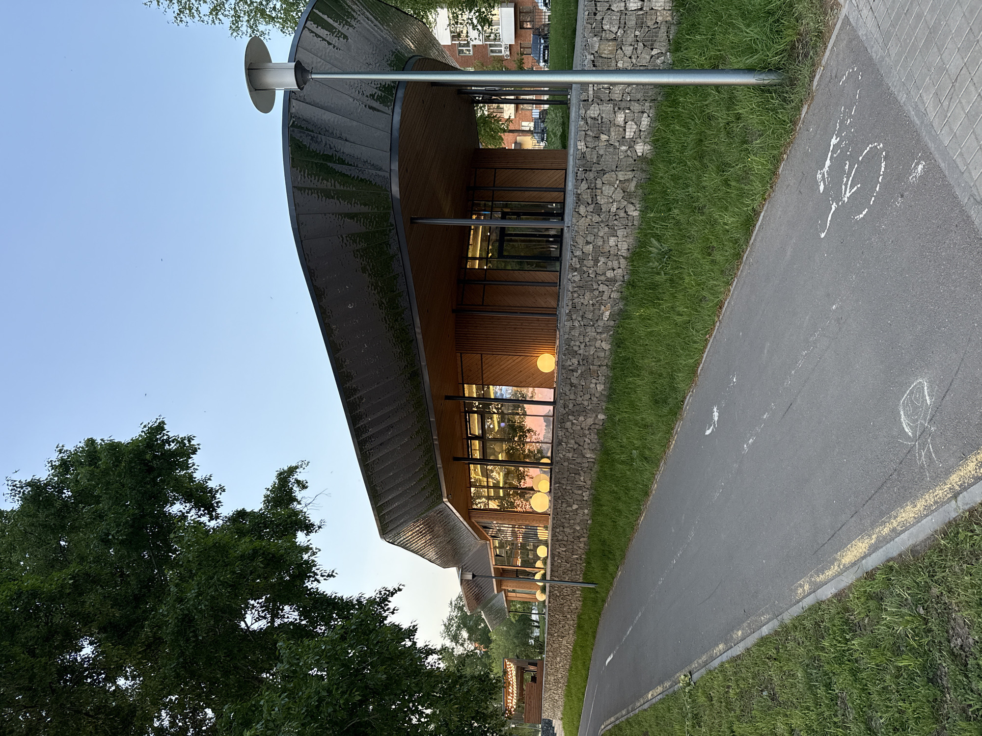 A view of a modern building in Votkinsk, Russia, features a dark, curved roof and large windows. The building has stone walls and is bordered by a grassy area and a paved path. A street lamp is visible in the foreground. The path has painted bike lane markings. Trees are visible in the background.