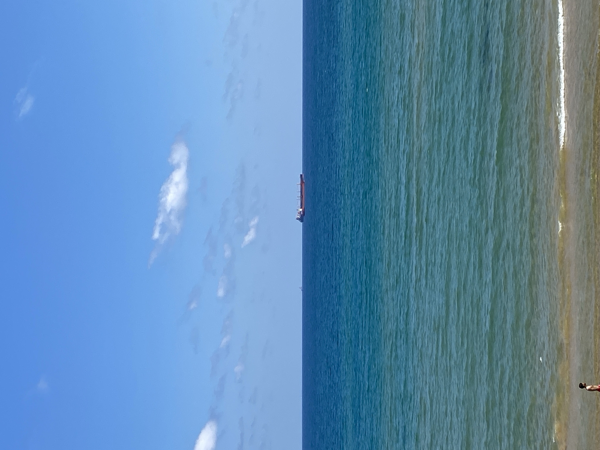 A wide shot captures a tranquil beach scene on a bright, sunny day with a clear blue sky dotted with a few white clouds. The vast expanse of the ocean dominates the frame, with two distinct shades of blue indicating different depths and the play of light on the water. The closer waters are a vibrant turquoise, transitioning to a deep, rich blue in the distance. A large cargo ship, painted in shades of red and white, sails horizontally across the middle of the ocean, a solitary vessel on the horizon. On the sandy shore, a lone person, appearing to be a male, walks along the water's edge. The beach itself is a strip of light brown sand, with gentle waves lapping at the shore, creating a thin line of white foam. The scene is serene and evokes a sense of peace and openness, likely depicting a location along the coastline of Sarıyer, Turkey, a district known for its beautiful Bosphorus views and seaside resorts.
