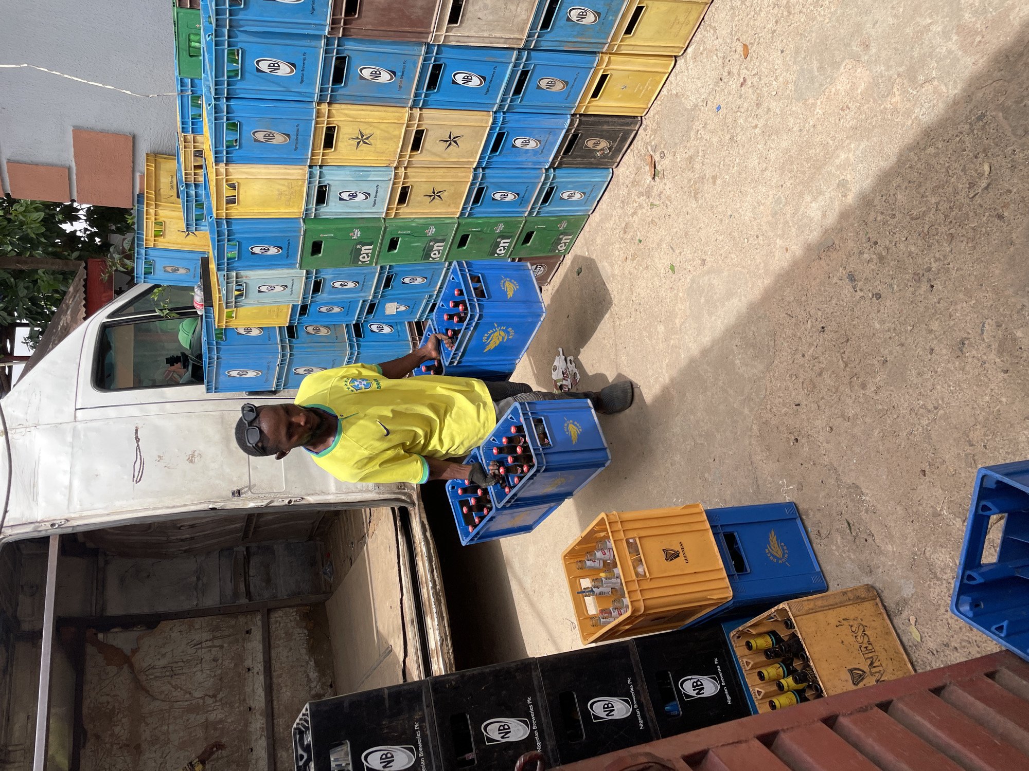 This media file captures an outdoor scene in Jos, Nigeria, depicting a man engaged in the manual labor of transporting beverage crates. The setting appears to be a commercial or distribution area, likely a loading bay or storage facility, characterized by stacks of plastic crates and a white van.
The central subject is a dark-skinned man, wearing a bright yellow t-shirt with a Brazilian flag crest and a Nike logo, black trousers, and dark boots. He also wears dark sunglasses on his head. He is actively carrying two blue plastic crates, each filled with multiple bottles, possibly beer or soft drinks. One of the crates he holds has "PREMIUM BREW" printed on it. He appears to be loading or unloading these crates from the rear of the white van, which has its back doors open, revealing a sparse interior.
The background and foreground are dominated by numerous stacks of beverage crates of various colors – blue, yellow, green, and black. These crates are branded with logos and text, including "NB" and "Nigerian Breweries Plc" on many blue and black crates, "Heineken" (or "ken") on green crates, and "Guinness" (or "NINESS!") on some yellow and black crates. Some crates are empty, while others contain bottles.
The ground is concrete, showing some dirt and debris. Strong, sharp shadows cast across the concrete indicate bright daylight, suggesting it is likely midday or early afternoon. The overall impression is one of active commerce and distribution in a bustling environment.