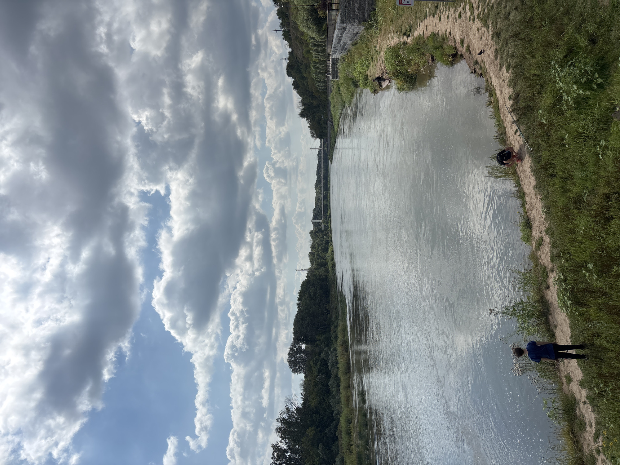 The image depicts a wide river with a gentle current, flowing through a landscape characterized by lush green vegetation and a mix of deciduous trees lining the banks. The sky is a dynamic canvas of bright blue interspersed with large, fluffy white and grey clouds, suggesting a partly cloudy day.
In the foreground, along the sandy riverbank, two young males are present. One, wearing a dark blue t-shirt and black pants, stands facing the river, holding a long, thin object that appears to be a fishing rod. The other individual, in a black t-shirt and shorts, is crouched down near the water's edge, seemingly engaged in an activity near the river. A dark-colored bird is also visible on the bank.
The river extends towards a distant horizon where a suspension bridge is clearly visible. The bridge spans the river, connecting the two banks. The surrounding terrain is hilly, with trees and foliage covering the slopes. On the right bank, a paved walkway with a railing runs along the edge, interspersed with trees. A stone wall supports this elevated area. In the far distance, on the right bank, a telecommunications tower can be observed.
The scene is consistent with the embankment of the Ural River in the city of Orenburg, Russia, a location known for its scenic river views and recreational spaces. The overall atmosphere is tranquil and suggests a peaceful outdoor setting.