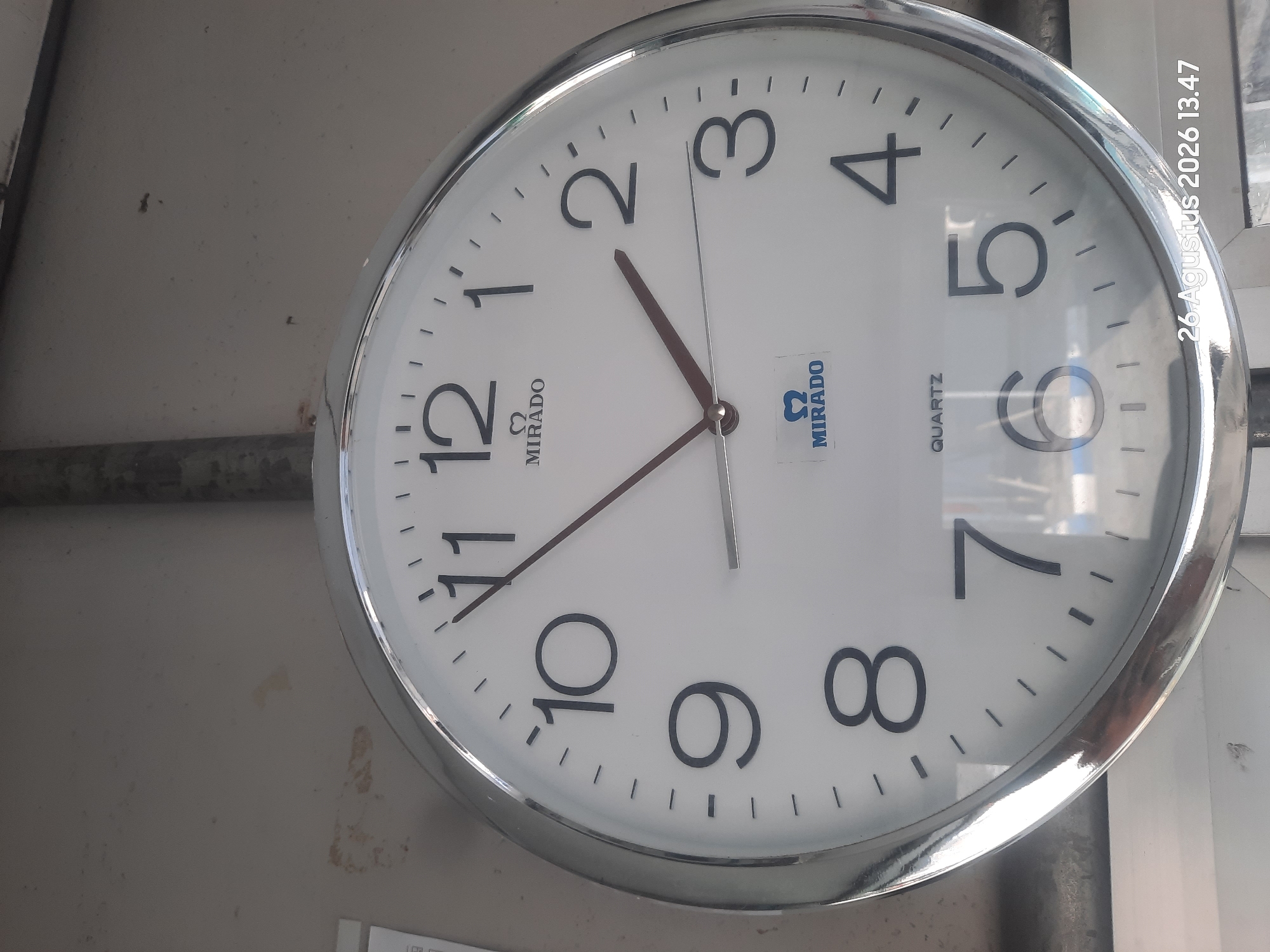 A close-up, slightly angled shot shows a white analog clock with black Arabic numerals and markers. The clock face has two dark-colored hands: an hour hand and a minute hand. The hour hand points slightly past the "1" and the minute hand points slightly past the "2", indicating the time is approximately 1:11. The clock also displays the brand "MIRADO" in black text below the "12" numeral and a "MIRADO" logo with a stylized "Ω" symbol below the "6" numeral. The word "QUARTZ" is printed in smaller black text below the "6". The clock is framed by a reflective silver bezel. In the bottom right corner of the image, a timestamp is visible: "26 Agustus 2026 13.47". The background consists of a neutral, textured wall and a vertical metallic pipe. This image was captured in Gending, Indonesia.