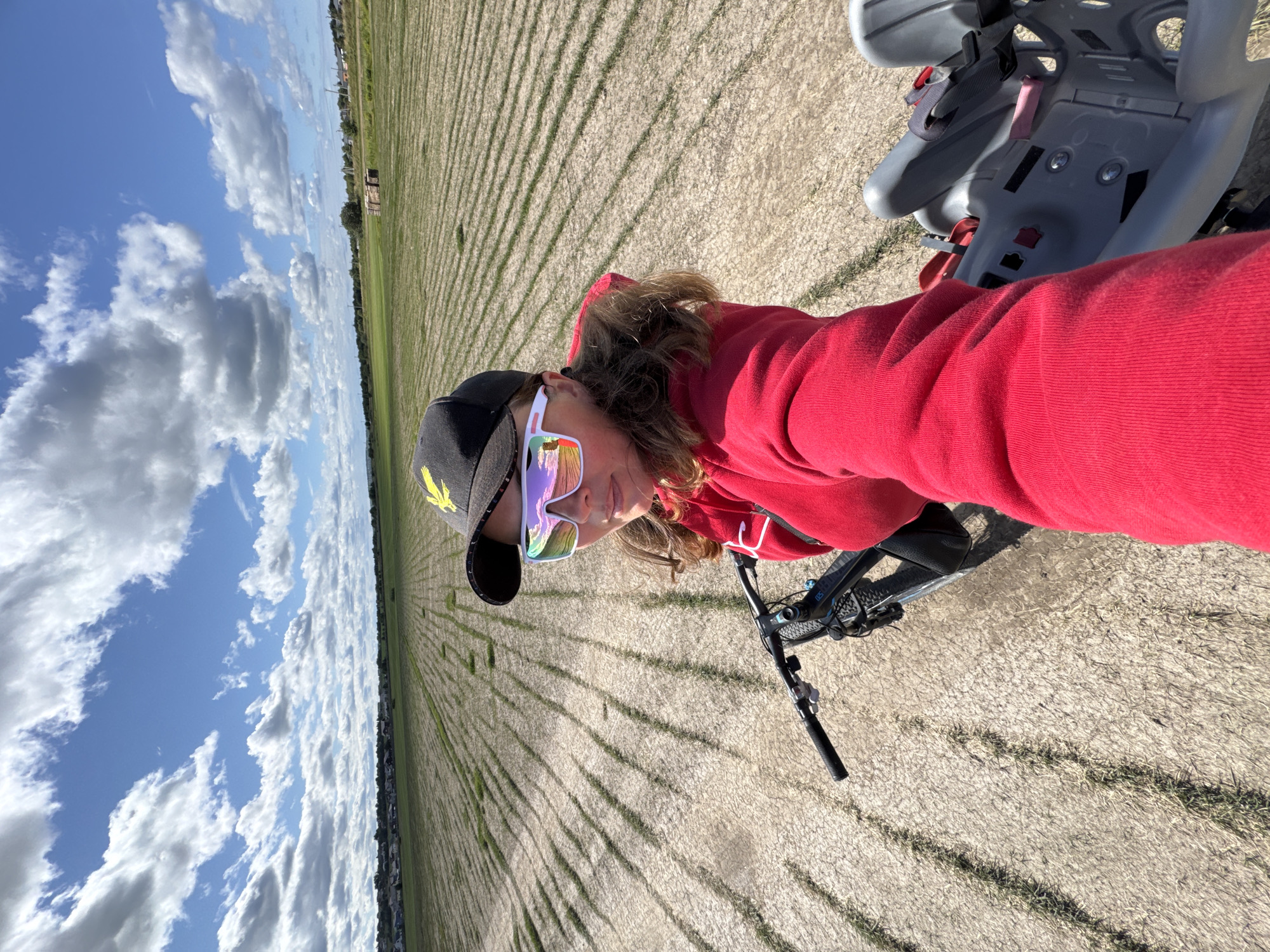 An outdoor, high-angle, wide shot shows a woman riding a bicycle in a field on a sunny day. The woman, wearing a red hoodie, a black baseball cap with a yellow eagle logo, and white mirrored sunglasses, is in the foreground and is looking at the camera. The bicycle has a grey child seat attached to the back. The field behind her is dry and has visible rows of what appears to be stubble or dried plants. In the background, a line of trees separates the field from a residential area with houses and a few taller structures. The sky is blue with scattered white clouds. The location is the city of Kempele, Russia.
