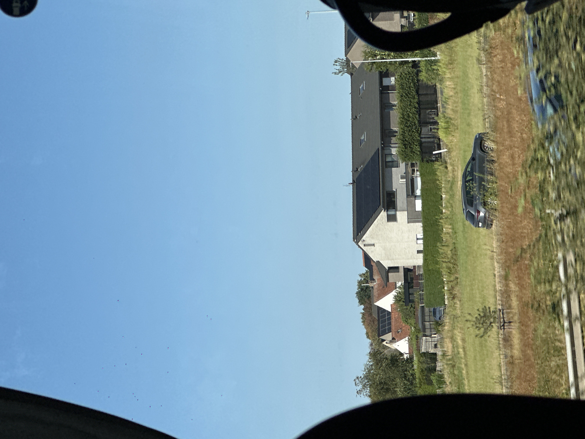 The image captures a view of a residential area in Oostkamp, Belgium, on a clear, sunny day. The foreground is dominated by the interior of a vehicle, with a dark curved frame on the left and right sides of the image suggesting windows and potentially part of the vehicle's structure.
In the mid-ground, a road runs horizontally across the frame, with a gray SUV driving on it. Adjacent to the road is a grassy field with scattered dry vegetation and a small, sparse tree. Beyond the field, a row of modern houses is visible. These houses are characterized by dark gray tiled roofs, some of which are fitted with solar panels. The houses have light-colored facades, and some feature dark-framed windows and doors. A dense, neatly trimmed green hedge runs along the front of some of the properties. In the background, more trees and houses can be seen, with a clear blue sky overhead.
There are no discernible people in the image. The overall scene depicts a suburban or semi-rural environment, typical of many residential areas.
