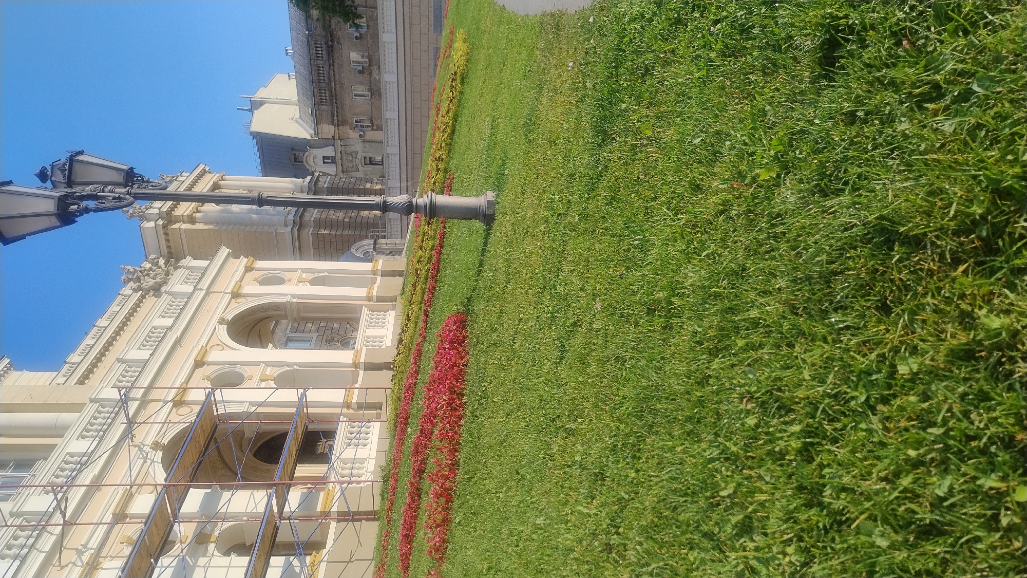 A low-angle, medium shot captures a portion of the exterior of a grand, light-colored building, possibly an opera house or theater, with scaffolding on its left side. The building features ornate architectural details, including arched entrances and decorative carvings. In the foreground, a lush green lawn slopes upwards, with beds of red and yellow flowers arranged in rows along the incline. A dark, antique-style lamppost stands prominently on the right side of the lawn. Behind the main building, other structures with grey roofs are visible, suggesting an urban setting under a clear blue sky. The scene appears to be set outdoors during daylight hours. There are no people or animals in the frame.
