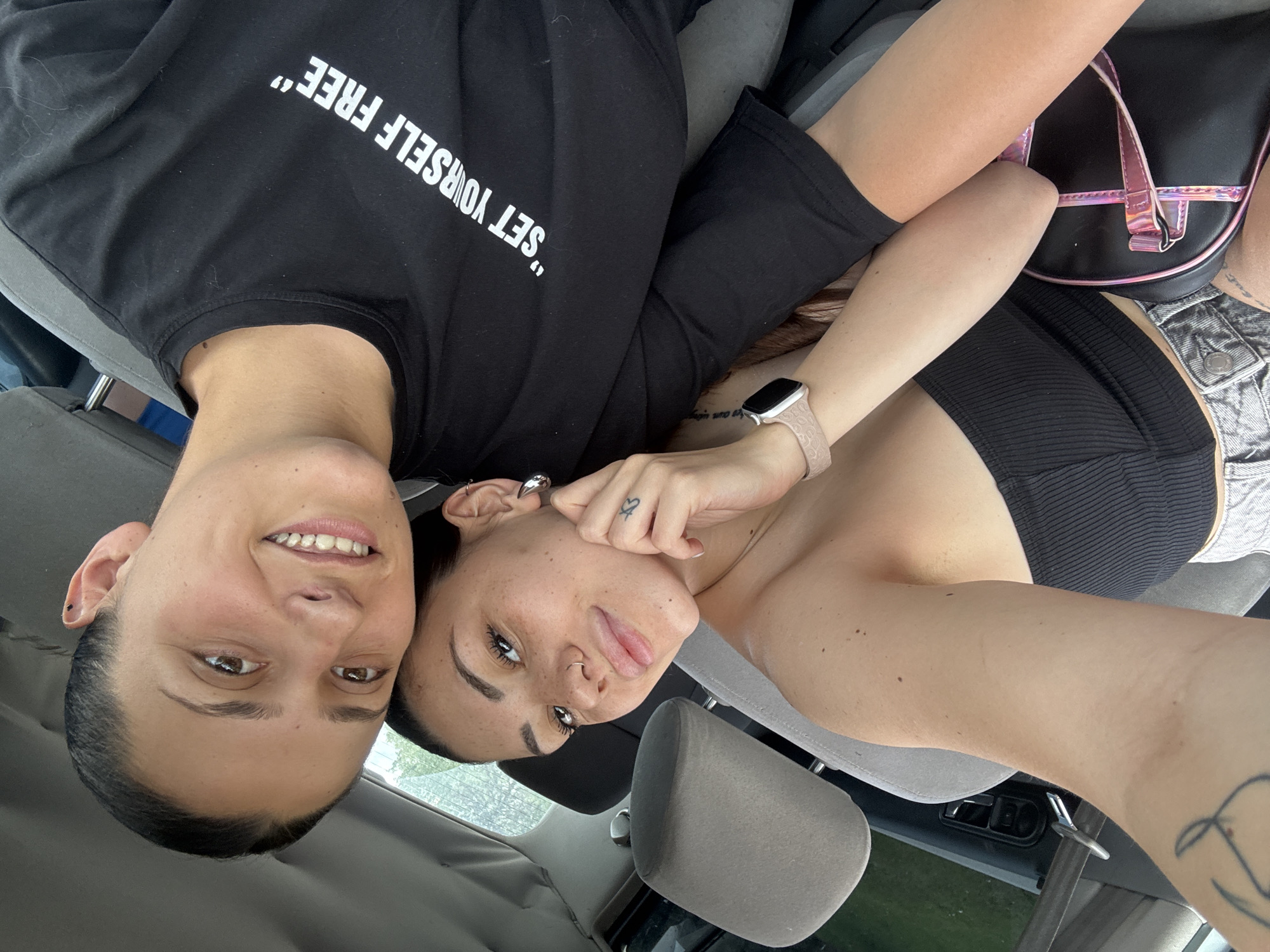 The image displays two young adult females inside the front passenger compartment of a car. The location cannot be confirmed as Sofia, Bulgaria, or any specific geographic area based on the visual evidence.
The female on the left, positioned in the foreground and appearing to take the selfie, has light skin, dark hair pulled back, and a face with visible freckles. She exhibits a septum piercing, a nostril stud, and large, silver teardrop-shaped earrings. Her attire consists of a black ribbed crop top, with the waistband of light-colored, possibly denim, shorts visible on her lap. A light-colored Apple Watch with a textured band is present on her right wrist, and a small heart tattoo is visible on her inner right wrist. A faint, abstract tattoo is also visible on her upper left arm. She maintains a slight smile while looking directly at the camera, with her right hand resting near her chin.
The female on the right has light skin, dark hair pulled back, and is wearing a black crew-neck t-shirt. The white text "SET YOURSELF FREE" is prominently displayed on her shirt. A small black stud earring is visible in her left ear. She is smiling broadly and also looking directly at the camera.
The car interior features grey fabric seats and headrests. A dark door panel with a silver door handle and lock mechanism is visible on the left. A seatbelt buckle is also visible. A black bag with iridescent pink straps and trim is situated between the two individuals, resting on the lap of the female on the left. The blurred background visible through the car windows indicates an outdoor, daytime setting with greenery.