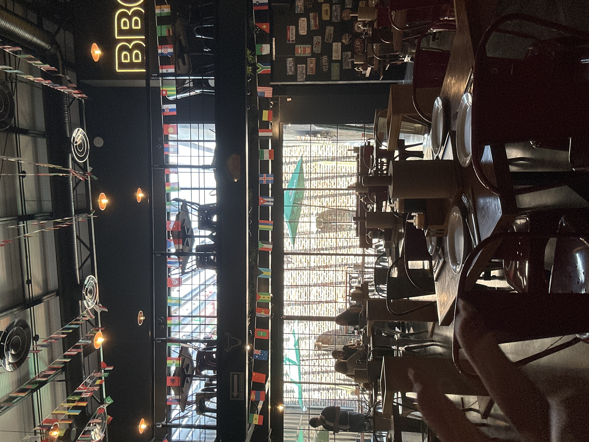The interior of a BBQ restaurant, possibly located in the city of Unknown, Unknown. The scene features rows of tables and chairs occupied by patrons. International flags hang from the ceiling alongside an illuminated "BBQ" sign. The walls are adorned with license plates. Natural light enters through a large window, illuminating the space.