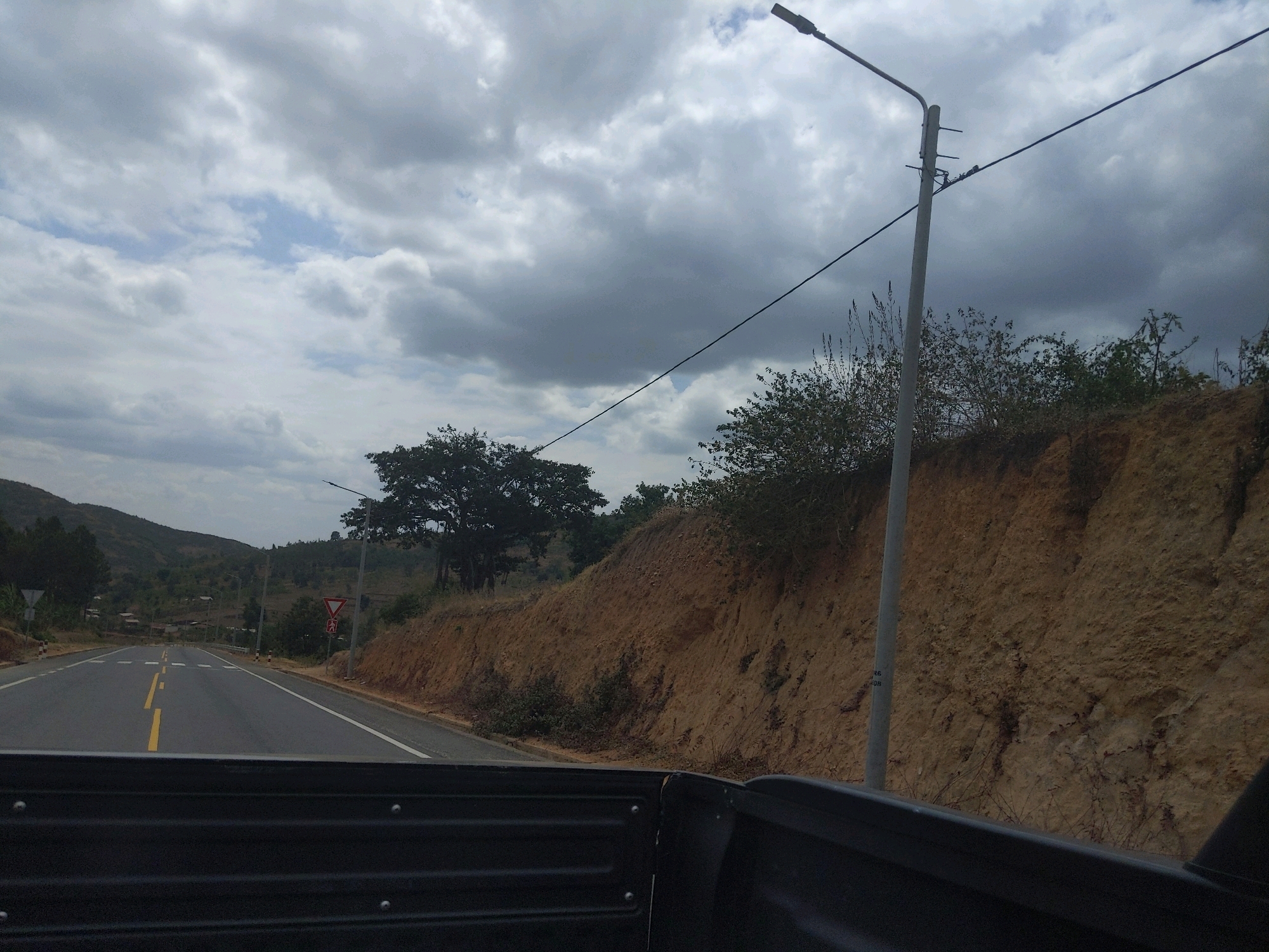 This image shows a view from the back of a vehicle traveling on a paved road in the city of Muyira, Rwanda. The road is lined with yellow and white markings. On the right side of the road, there is a steep, eroded dirt embankment covered with sparse vegetation. A tall lamppost with overhead wires stands on this embankment. Further down the road, there are more lampposts and a yield sign with a pedestrian symbol. The landscape beyond the road is hilly and covered with trees and some scattered buildings. The sky is overcast with dark clouds. The foreground is dominated by the dark, metallic interior of the vehicle's cargo area.