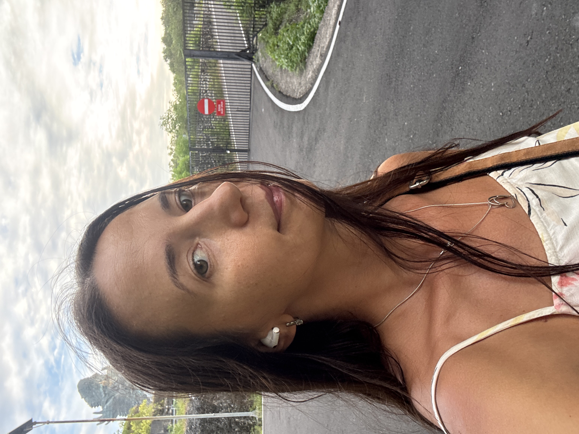 A woman with long brown hair, wearing a white floral-patterned top and a white earbud in her left ear, is posing for a selfie outdoors. She is wearing a necklace. Behind her is a paved road with a white border and a gate with a red and white circular sign. Vegetation is visible on the other side of the gate. The sky is overcast. Based on the visual features present, the location is in Kuta Selatan, Indonesia.