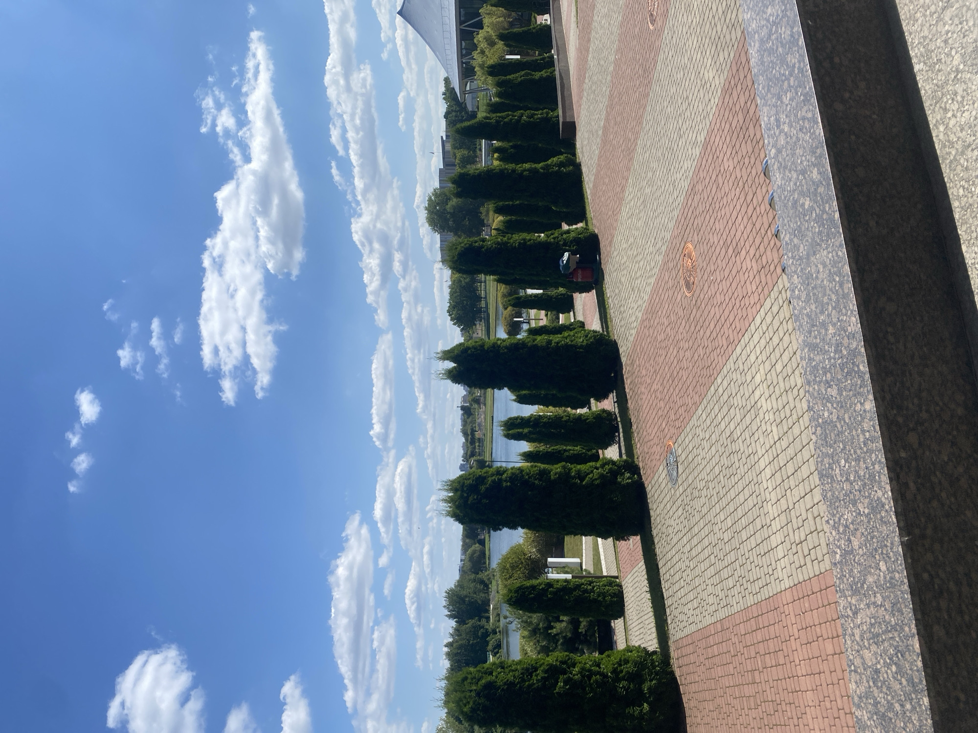 The image captures an outdoor scene on a sunny day with a bright blue sky filled with scattered white clouds. In the foreground, a paved area made of red and beige interlocking bricks stretches out. A granite-like structure runs along the bottom of the image. Behind this paved area, a line of tall, dark green evergreen trees stands erect, creating a natural barrier.
Beyond the trees, a calm body of water, likely a lake or river, is visible, with a distant treeline and buildings on the far bank. On the right side of the image, a portion of a modern building with a large, sloping roof is present. In the middle ground, a red trash receptacle with a white top is situated next to one of the evergreen trees. The overall atmosphere suggests a well-maintained public space, possibly a park or a promenade area in a city.
This scene is located in the city of Minsk, Belarus, specifically at the Museum of the Great Patriotic War.
