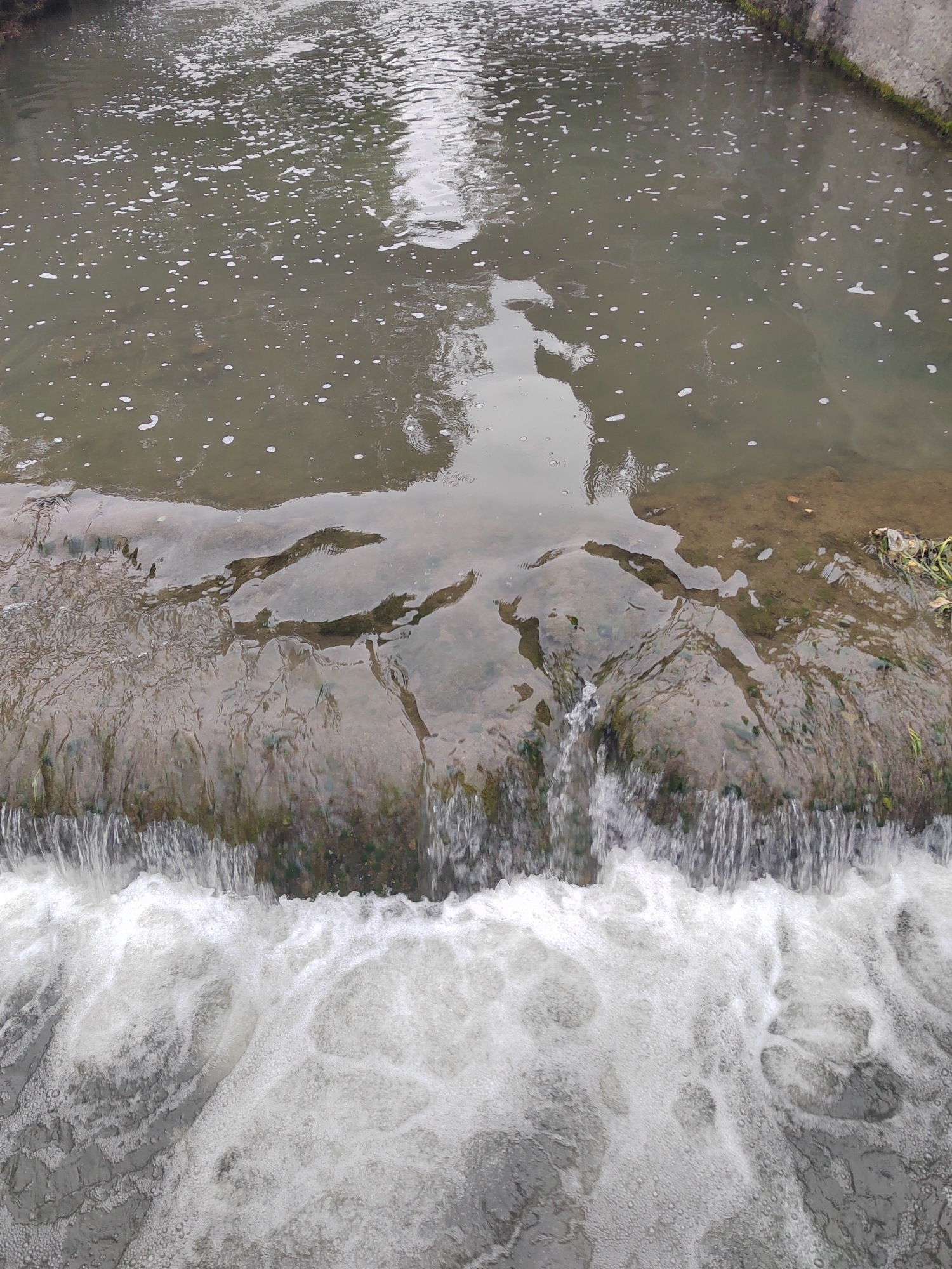 The image shows a small waterfall or spillway in a body of water. The water appears to be somewhat murky or turbid. A concrete or stone ledge is visible, over which the water cascades, creating a white foamy effect at the base. The water surface above the ledge has ripples and reflections. The environment seems to be natural, potentially within an urban or suburban setting, but specific location details are unknown. There are no people visible in the frame.