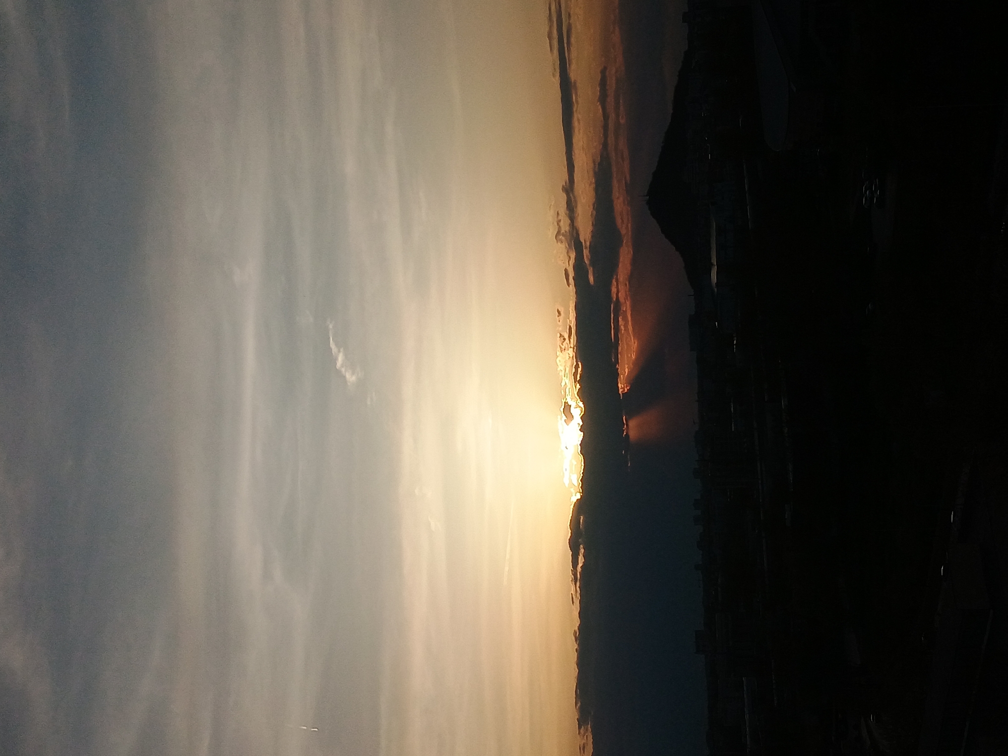 The image captures a sunset scene, likely from a vantage point in Plovdiv, Bulgaria. The sky transitions from a pale blue-gray on the left to a warm, golden-yellow and orange gradient near the horizon. The sun is partially obscured by clouds along the horizon line, creating rays of light that penetrate downwards. Silhouetted in the foreground, possibly a skyline or buildings, are dark, undefined shapes. The overall impression is that the photograph captures a moment of natural beauty at sunset, with an urban or built environment in the distance.
