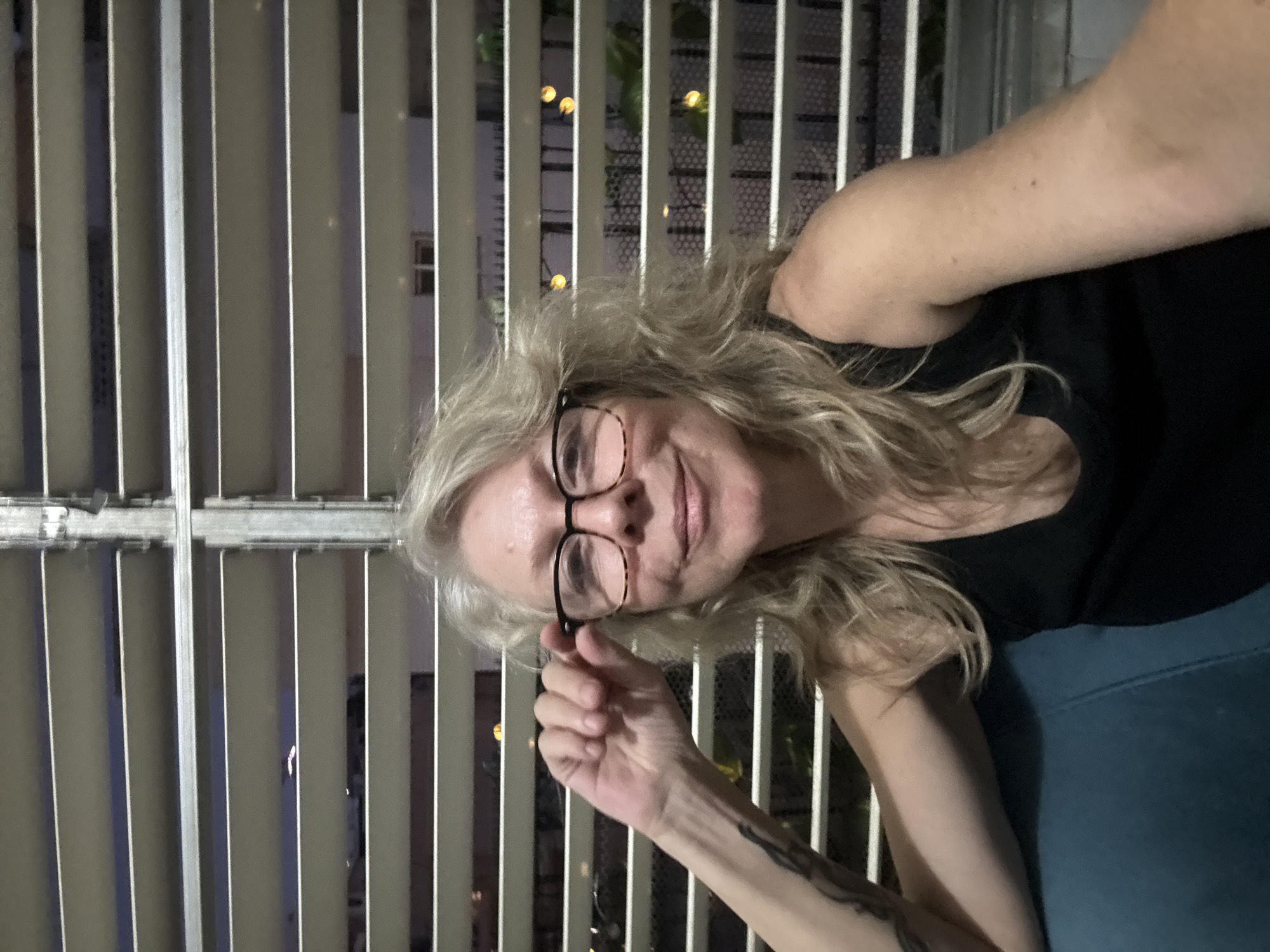A woman with graying blonde hair and glasses is sitting on a teal couch, looking at the camera and adjusting her glasses. She is wearing a black tank top. Behind her are horizontal blinds, with an outdoor scene visible through the gaps. The scene outside appears to be at night, with some lights and foliage visible. The environment suggests a residential setting, possibly an apartment in Ramat Gan, Israel.
