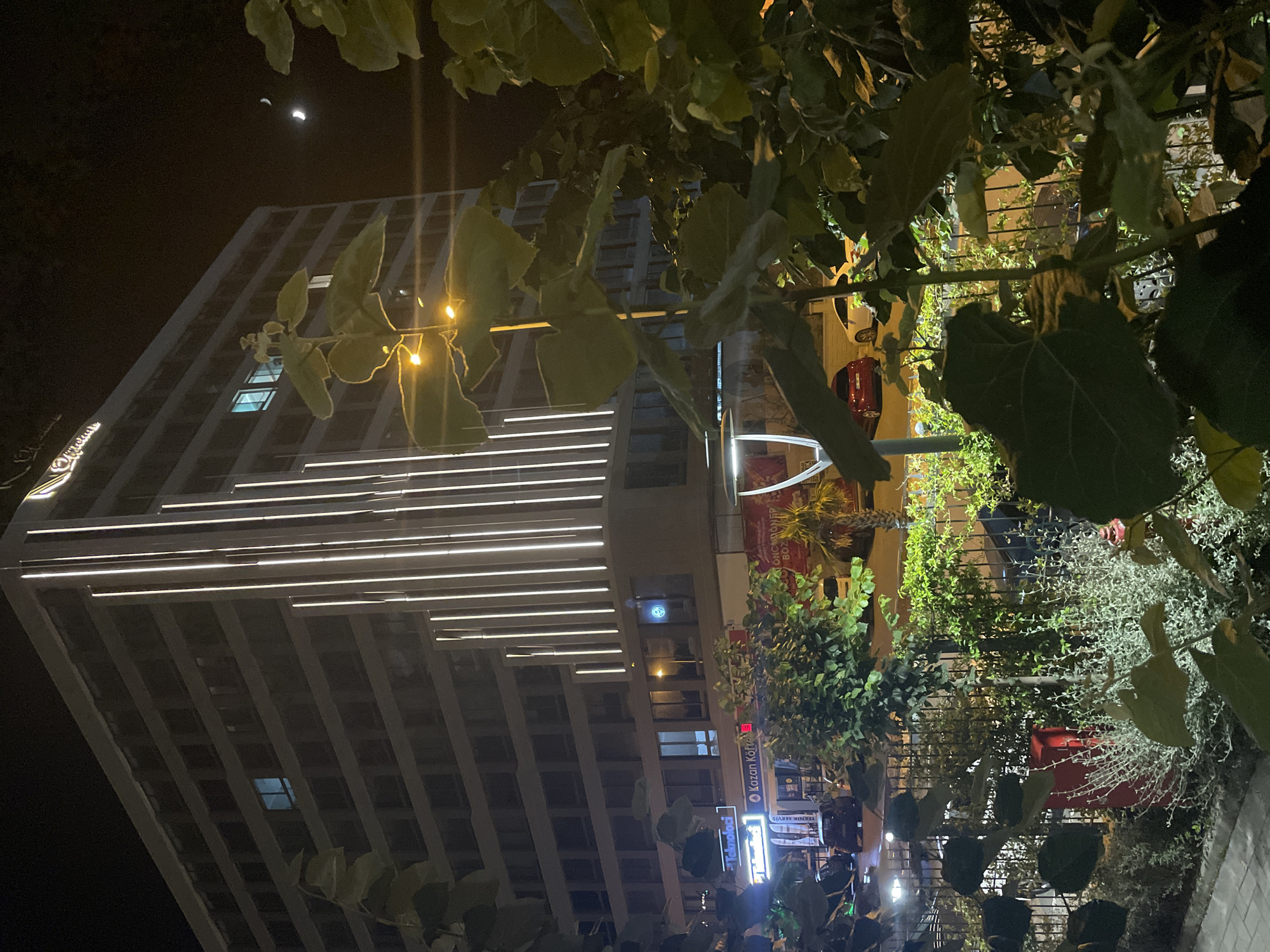 The image depicts a nighttime scene in the city of Yunus Emre, Turkey. A modern, multi-story building with vertical white lights illuminating its facade dominates the background. The building has numerous windows, some of which are illuminated from within. A sign with a logo and text, possibly a business name, is visible at the top of the building.
In the foreground, lush green foliage partially obscures the view of the building and the street below. Several large leaves are prominent in the lower portion of the image. Beyond the foliage, a fence separates the foreground from a sidewalk and street. Parked cars, including a red and a white vehicle, are visible on the street.
Additional elements include streetlights casting a yellow glow and two crescent moons visible in the dark night sky. Signs for businesses, including "Teknoloci" and "Kazan Köfte," are discernible on the lower levels of the building. The overall impression is a well-lit urban environment at night, with a blend of modern architecture and natural elements. No people are directly visible in the scene.