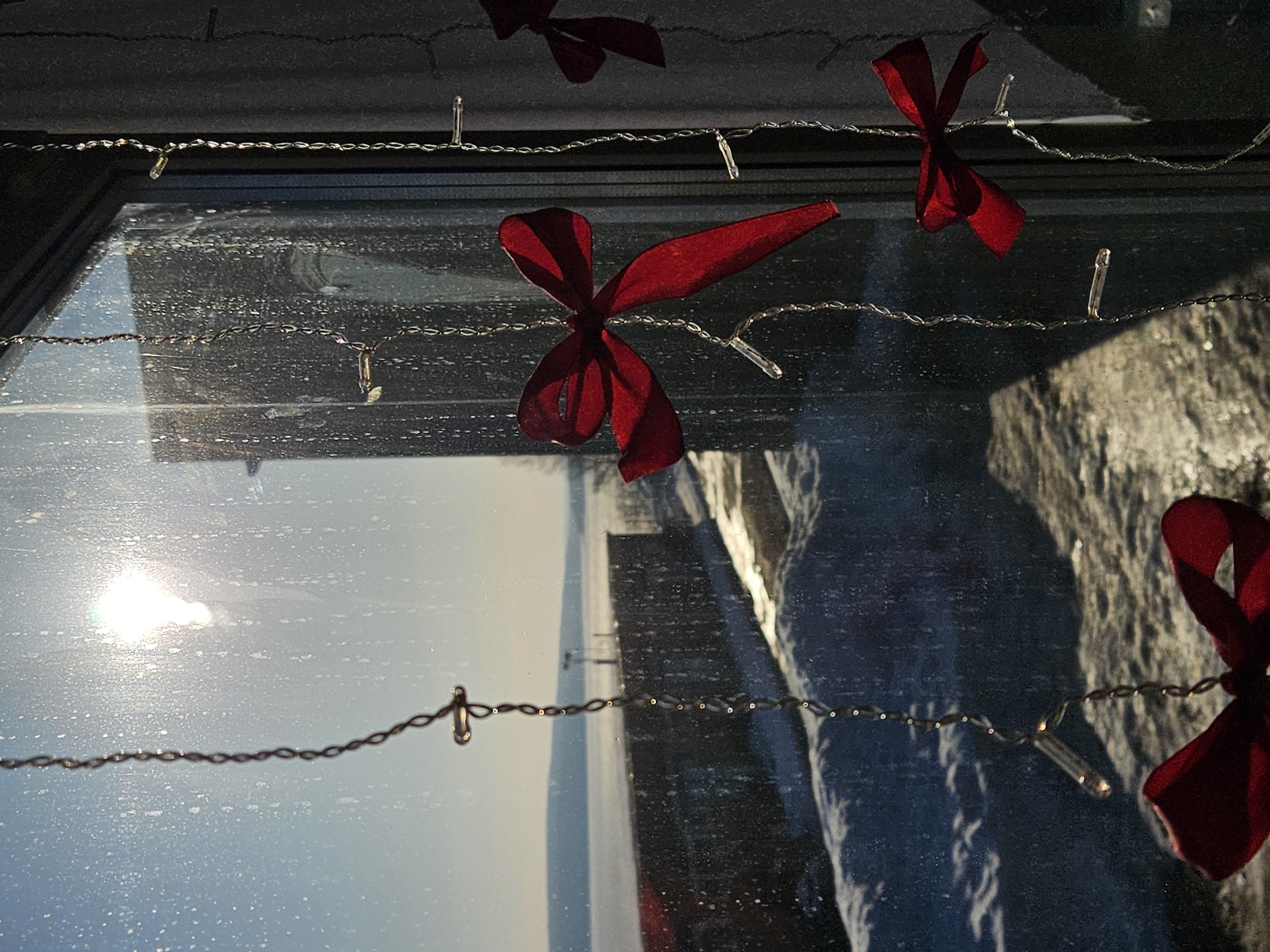 The image displays a detailed winter scene viewed from inside a building in Chaikovsky, Russia, looking out over the Votkinsk Reservoir. The foreground shows the interior side of a window, which is noticeably spotted or dirty, possibly with dust or water residue. Bright sunlight from the upper left creates a strong flare on the glass. Attached to the window, likely from above, are several decorative strands consisting of thin, silver-colored chains. These chains are adorned with small, clear, cylindrical ornaments and at least three red velvet bows: a large one positioned in the mid-right, a smaller one at the bottom-center, and a partial one visible at the top-right.
Through the window, the outdoor environment is visible. The ground is covered in uneven snowdrifts. A dark, rectangular building with a flat roof stands in the midground, accompanied by a smaller red structure to its left. Beyond these structures, the expansive Votkinsk Reservoir appears frozen and covered in snow or ice, extending towards a distant, undulating horizon of hills or landmasses under a clear, pale blue sky. Sunlight illuminates the snowy landscape, causing reflections on the ground and ice. The dark frame of the window is visible along the right edge and top.