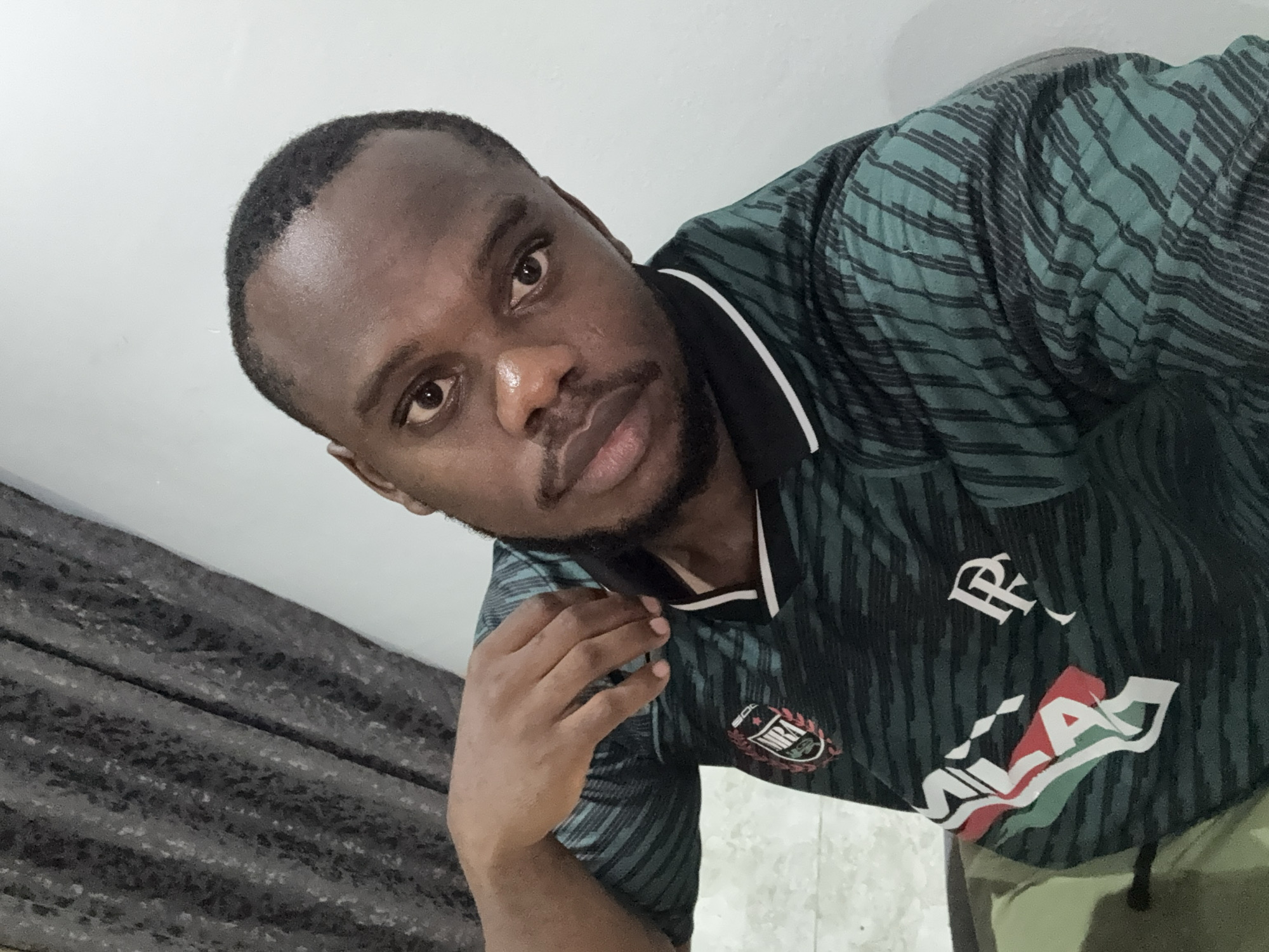 An adult man with dark skin, short dark hair, and a short beard takes a selfie in what appears to be an indoor setting in Iddo Tudunwada, Nigeria. He looks directly at the camera with a neutral to slightly serious expression. His right arm is bent, with his hand resting near his jawline. He is wearing a dark green polo shirt patterned with black diagonal stripes, featuring a black collar with a white stripe. Several logos are visible on the shirt: a circular emblem with the word "MIRA" on his left chest, a white stylized letter "R" on his right chest, and below it, a prominent logo displaying "SALA" in white lettering over a red, white, and green design. The background consists of a plain light-colored wall or ceiling above his head and to his left, and a dark, textured curtain or fabric behind him and to his right. The lighting is soft and appears to be artificial, suggesting an indoor shot, possibly in the evening or a room with closed curtains.