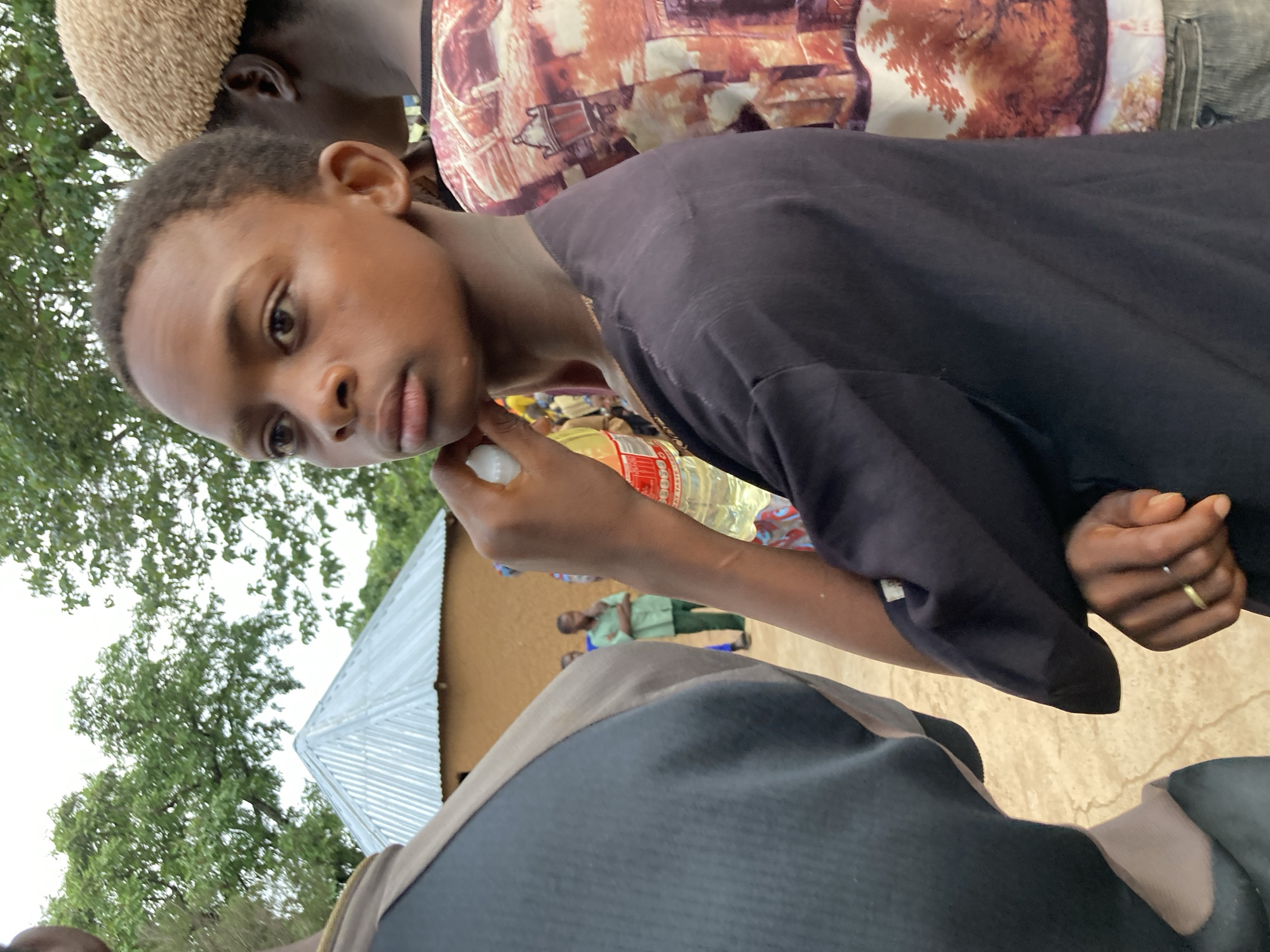 A young boy, appearing to be of African descent, stands in the foreground, looking to his right. He is wearing a dark, possibly black, short-sleeved shirt. His right hand is raised to his face, holding a white object, possibly a bottle cap or a piece of candy. A thin gold ring is visible on the ring finger of his left hand. Behind him, to his right, another person is partially visible, wearing a light-colored, textured cap and a patterned shirt with orange and brown hues. To the left of the boy, another person is seen from the back, wearing a dark garment with a grey shoulder area. In the background, a simple, tan-colored building with a silver, pitched roof is visible, surrounded by trees. A few other individuals are faintly discernible in the background. The ground appears to be dirt or sand. The overall setting suggests an outdoor gathering in a rural or semi-rural area. The image was taken in Jos, Nigeria.