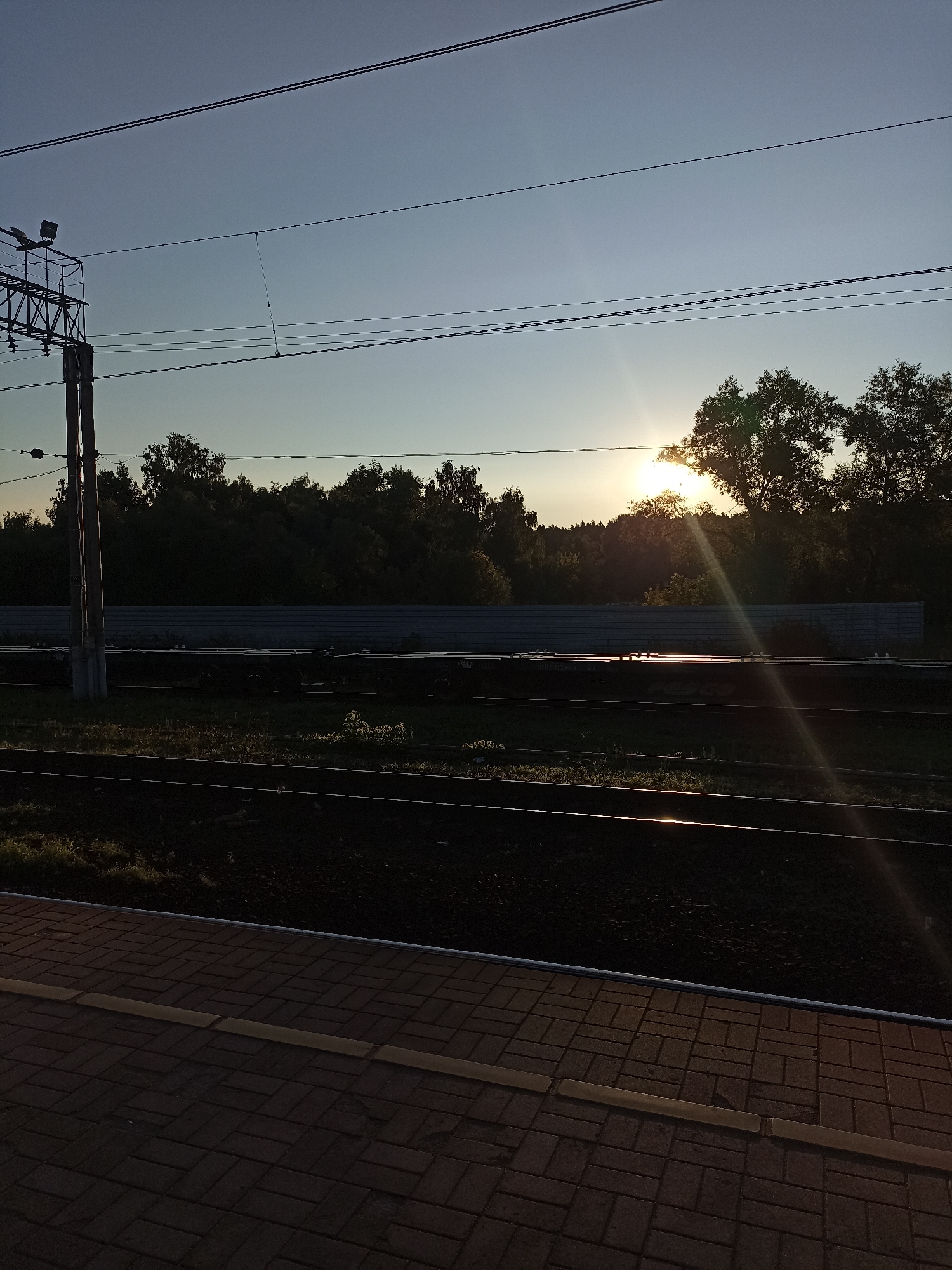 The image depicts a sunset scene at a railway station. In the foreground, there is a paved platform with yellow and brown brickwork. Adjacent to the platform are two sets of train tracks, with the tracks and surrounding area cast in shadow. Beyond the tracks, a dark, low wall runs horizontally across the frame. A dense line of trees forms the background, with the sun setting behind them, casting warm light and rays. Overhead, there are multiple power lines supported by a tall, metal pole with a lamp attached. The sky is a clear, pale blue, transitioning to a deeper blue towards the top. The overall impression is one of a serene, late-evening atmosphere at a quiet railway station.