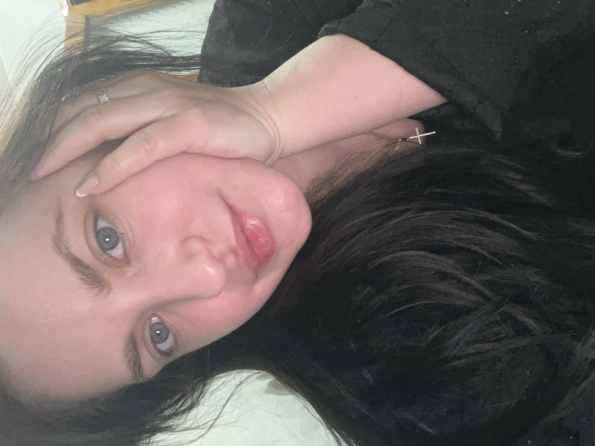 A close-up portrait shows a person, likely a woman, with dark hair, fair skin, and light blue-grey eyes, looking directly at the camera. The image is rotated, suggesting the subject is lying down with her head tilted. Her expression is soft, with a slight, subtle smile on her lips. Her right hand is resting gently beside her face, revealing a silver-toned ring with small sparkling elements on her ring finger and French-tipped fingernails. A delicate necklace with a small, sparkling cross pendant is visible around her neck. She is wearing a dark-colored top, possibly black. The background is blurred and indistinct, indicating an indoor setting, possibly a bed or comfortable surface. No specific time of day or weather conditions are discernible, and there is no visible text. The overall impression is one of relaxation and calm. The location context is Марковский, Russia.
