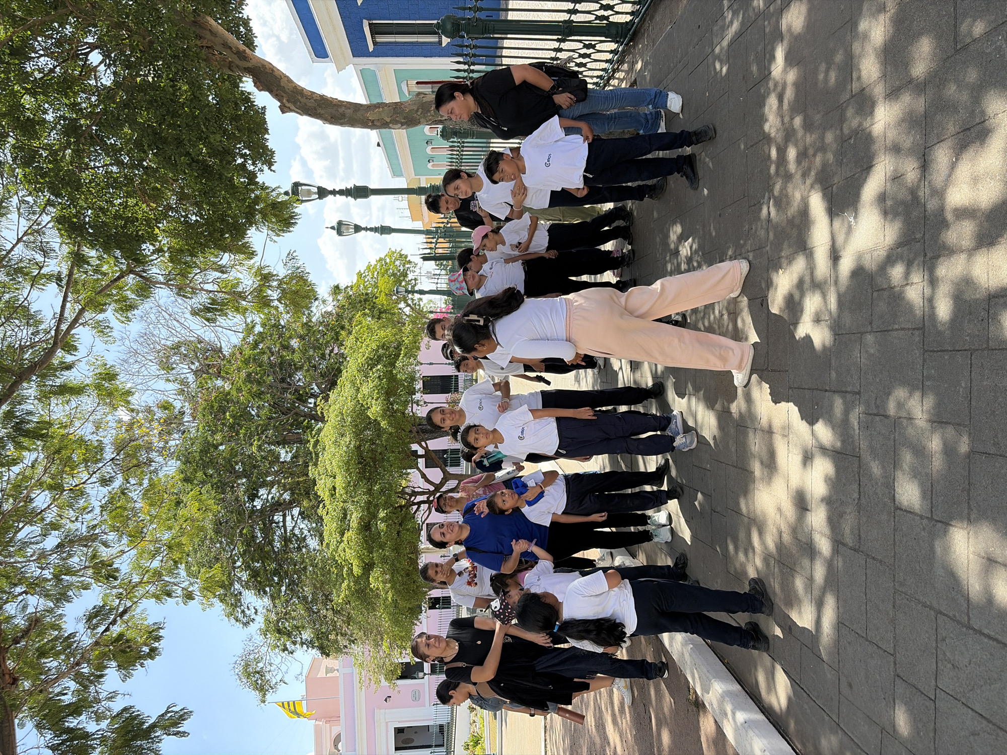 A group of about 15 people stand in a line on a paved sidewalk in Ciudad Bolivar, Venezuela. Most are young people wearing white shirts and dark pants or skirts. An adult woman in a black shirt and jeans stands at the end of the line. Trees with green foliage shade the sidewalk and line the street. Buildings in various colors such as pink, blue, and white are in the background. Streetlights are also visible behind the group.