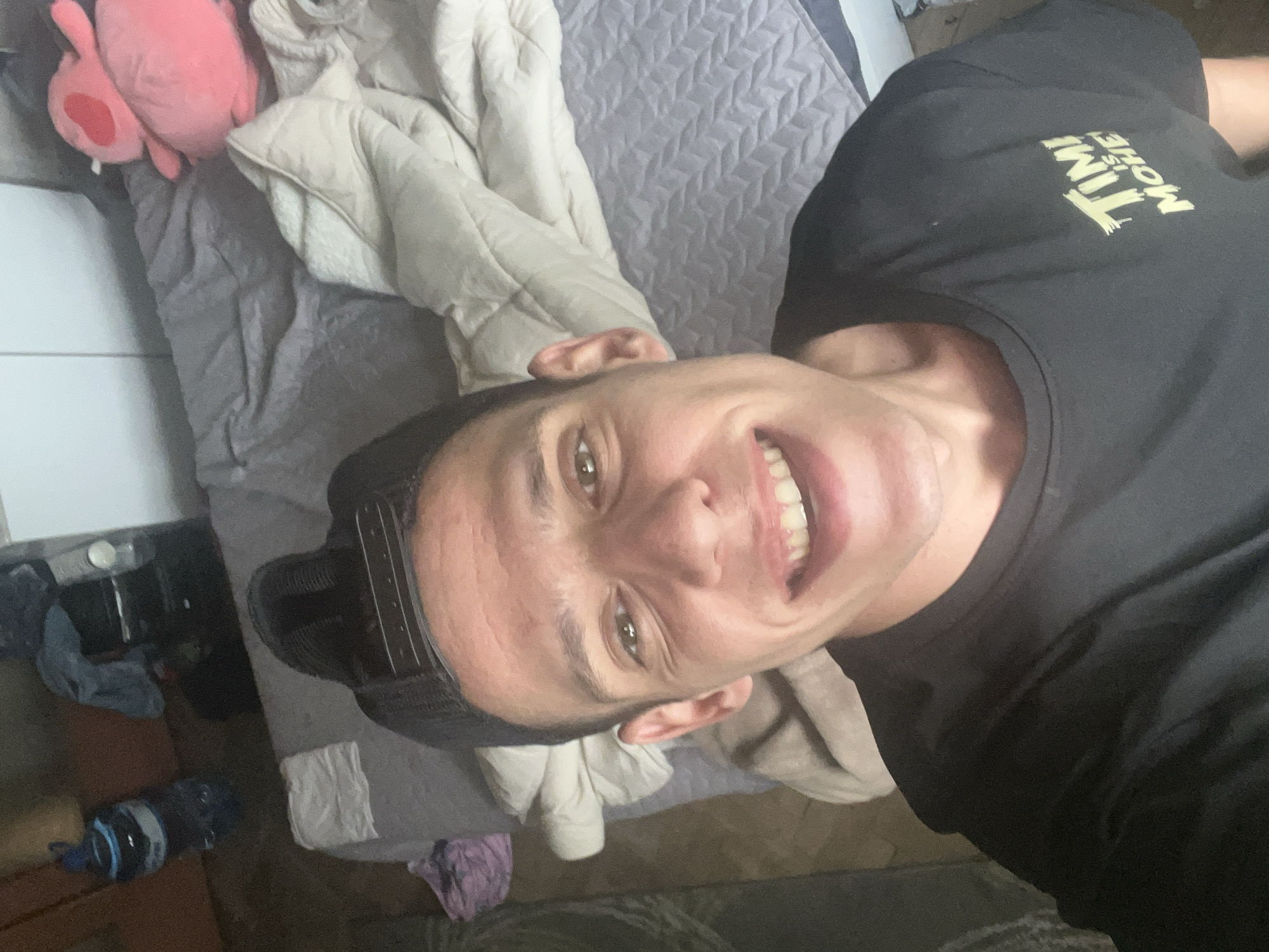 The image is a rotated selfie showing a young adult male in an indoor setting, likely a bedroom. The man is positioned in the foreground, smiling broadly at the camera with his teeth visible. He has short dark hair and dark eyes. He is wearing a black t-shirt and a dark baseball cap backward. On his t-shirt, yellow text is clearly visible, spelling out "TIME IS MONEY".
The background features a bed covered with a light grey patterned comforter. A cream-colored jacket or hooded garment is draped over the comforter. To the upper left, a pink plush toy, resembling a character like Kirby, rests on the bed. In the bottom left, some indistinct objects, including a dark water bottle, are visible on what appears to be a floor or low surface next to the bed, alongside a segment of a white tiled wall or surface. The man's cheerful expression suggests a positive mood.