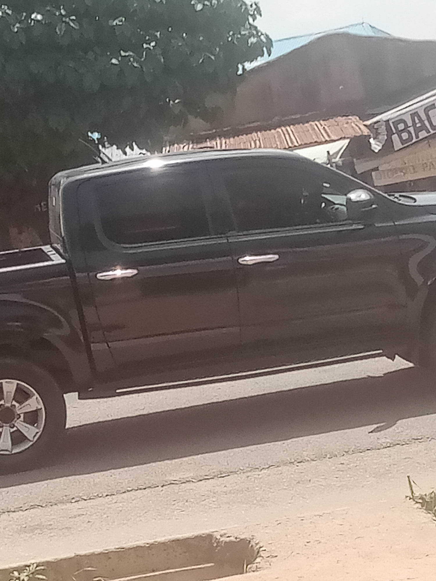 The image captures a side view of a black pickup truck parked on a street in Jalingo, Nigeria. The truck, with tinted windows and chrome door handles, is positioned at an angle, allowing a clear view of its passenger side. Behind the truck, a row of buildings with corrugated metal roofs is visible. One building has a sign with the partial word "BAC" in large letters. Trees with green foliage are present in the background, suggesting a moderately vegetated urban environment. The street appears to be paved, with a cracked surface indicating wear. The lighting suggests it is daytime, with a bright sky. The overall scene depicts a common street view in an urban setting in Jalingo.