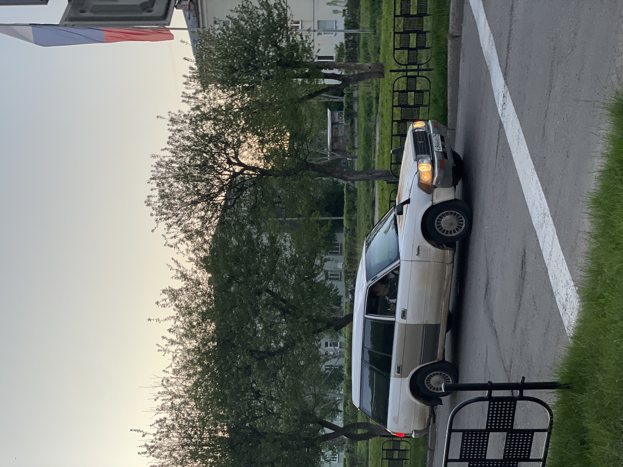 A white Toyota Crown is parked on the right side of a road, adjacent to a white line marking the edge of the pavement. The car's headlights are on. The license plate is visible and reads "K 844 TK 46". The vehicle appears to be occupied by an individual in the driver's seat. The background includes trees, grass, and buildings, characteristic of an urban environment. A structure with a Russian flag is visible in the top left corner. The location is Zheleznogorsk, Russia. Metal benches and other urban street fixtures are visible along the roadside.