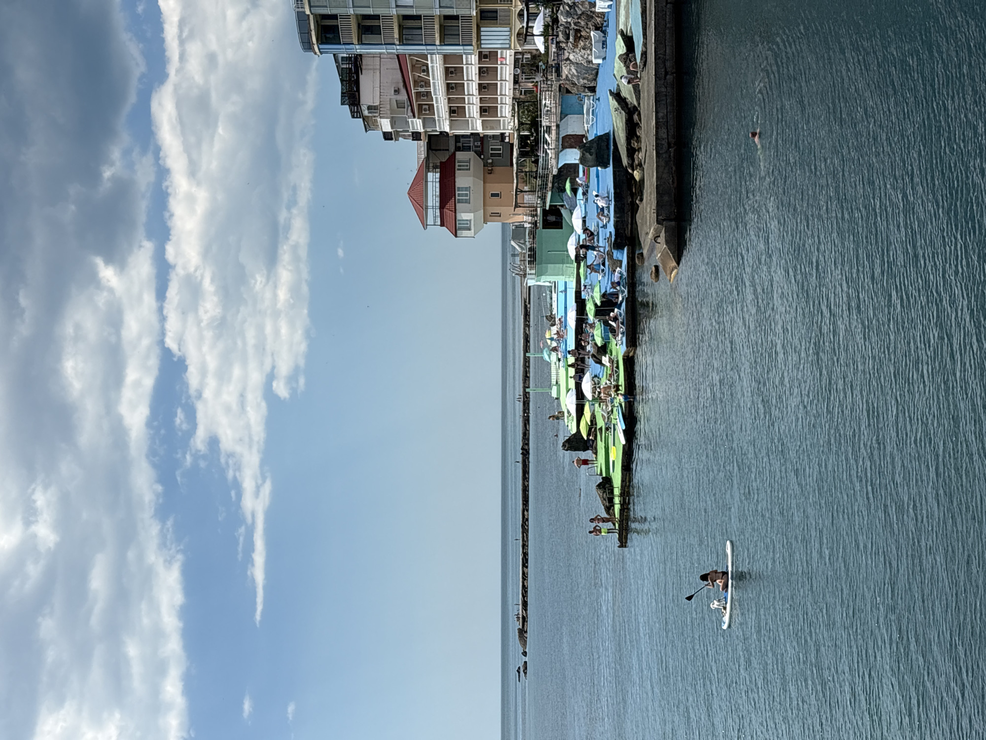 In Kiparisnoe, Ukraine, a picturesque coastal scene unfolds under a partly cloudy sky. The azure sea dominates the foreground, its surface rippling gently. A lone figure on a paddleboard, accompanied by a dog, glides across the water. In the mid-ground, a vibrant waterfront recreation area beckons with colorful inflatable structures, lounge chairs, and umbrellas. People are seen relaxing and enjoying the sun. The background is defined by a series of buildings lining the coast, their architecture reflecting a mix of styles. Further out, a long pier extends into the sea, with a few distant figures visible. The overall atmosphere is one of leisure and seaside tranquility.