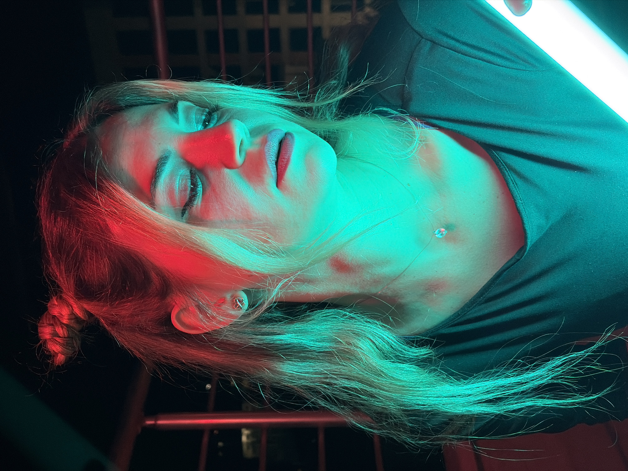 A young woman with blonde hair styled in a bun and loose strands is illuminated by red and teal lighting. She is wearing a dark-colored shirt and has on earrings and a necklace with a pendant. Her head is tilted down and her eyes are closed. The background appears to be a dark, outdoor setting with hints of urban structures. The image was taken in Budva, Montenegro.