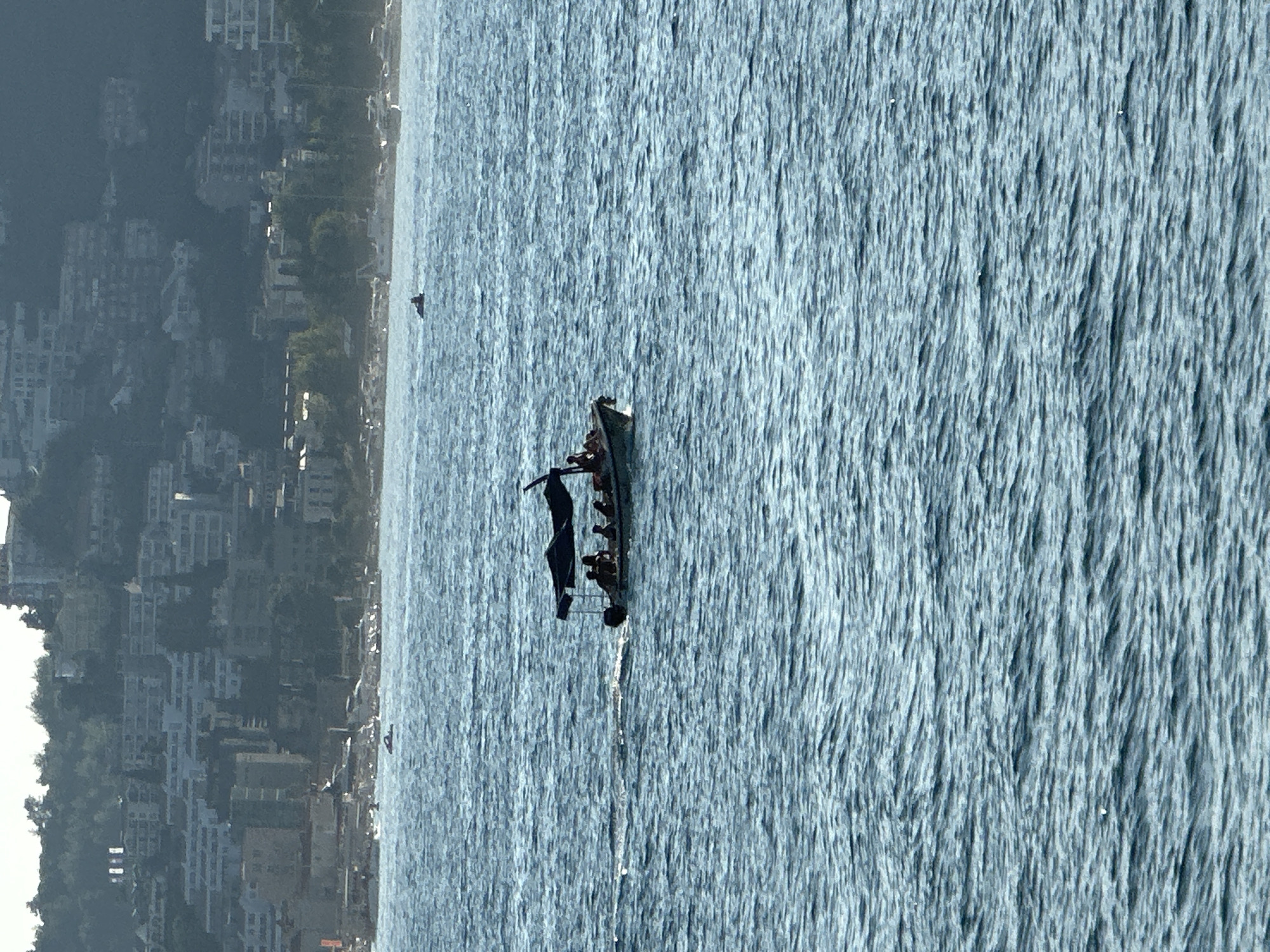 A speed boat with a dark canopy travels across choppy blue water. Six people are seated in the boat, appearing to be enjoying a ride. In the background, a hillside town with numerous buildings and dense foliage rises sharply. The town's architecture suggests a Mediterranean setting, likely along the coast of Budva, Montenegro, given the hilly terrain and densely packed, multi-story structures characteristic of the region. The water is the primary focus, with the distant town providing context for the scene.