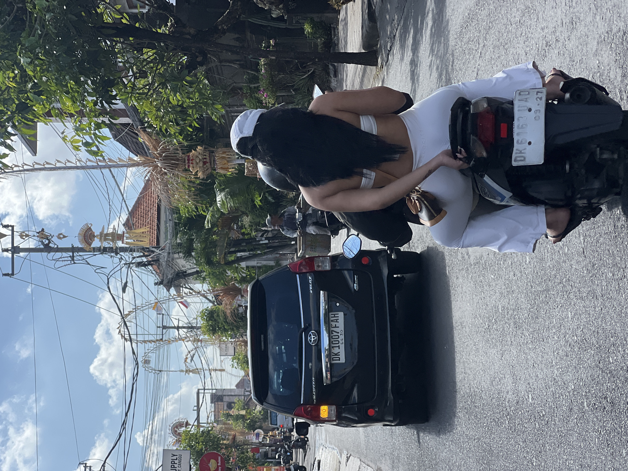 In North Kuta, Indonesia, a person rides a motorbike with a passenger behind them on a street lined with overhead wires and palm trees. The rider, wearing a white cap and black hair, is seen from behind, dressed in white athletic wear. The passenger, also in white, appears to be looking at a mobile phone. A black SUV with a license plate "DK 1007 FAH" is in front of them. The street is paved, and the sky is partly cloudy. Cultural decorations, possibly for a ceremony, are visible on poles and wires. Another motorbike with a rider is in the distance.