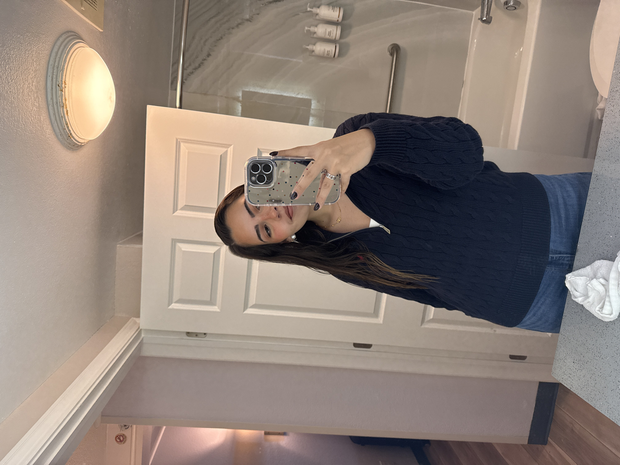 Here is a description of the image:
The image depicts a woman taking a selfie in what appears to be a bathroom. The bathroom has a white door, light fixture and a bathtub with bottles of shampoo. It is not possible to determine the exact location in San Antonio without additional information.