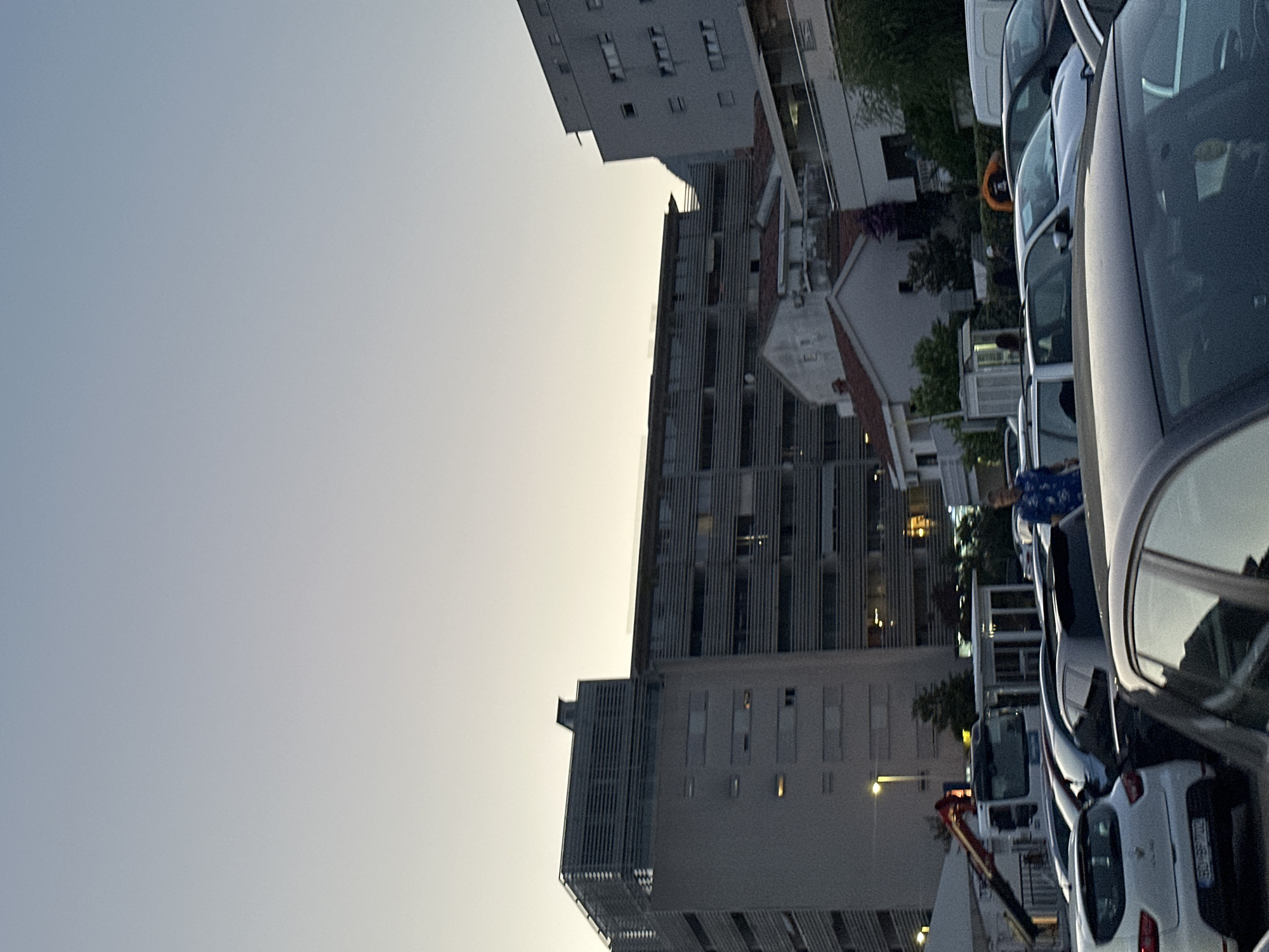 In Budva, Montenegro, a twilight scene unfolds in what appears to be a commercial or residential parking lot adjacent to several multi-story buildings. The sky is a gradient of pale blue to light yellow, suggesting either dawn or dusk.
Dominating the background are large, modern buildings with horizontal window lines and metallic louvers. One building, in the center-left, is a lighter gray, while the one to its right is darker and features illuminated windows. In front of these structures, smaller, older buildings with red-tiled roofs are visible, interspersed with trees.
The foreground is occupied by a variety of parked cars. A white Renault Clio is prominent on the left, its license plate readable. Several other vehicles, mostly sedans and hatchbacks, are parked in rows, some partially obscured. A white truck with a red crane arm is positioned on the left side of the frame, facing away from the viewer.
Several individuals are present. On the far right, a man wearing a blue patterned shirt stands near the parked cars, looking towards the left. Further into the scene, several other figures are visible in and around the parked vehicles, though their details are less distinct due to the distance and lighting. One individual appears to be walking near a small kiosk-like structure.
The environment suggests an urban setting, likely a developed area with mixed-use architecture. The lighting indicates a period of low natural light, characteristic of transitional times of day.